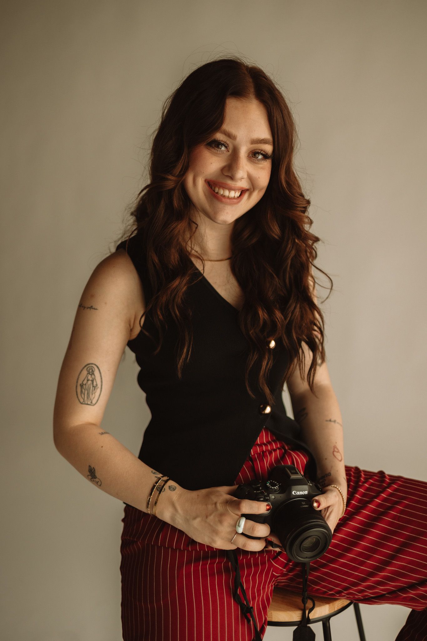 Person with long wavy dark hair holding a camera, wearing a black tank top and red striped pants.