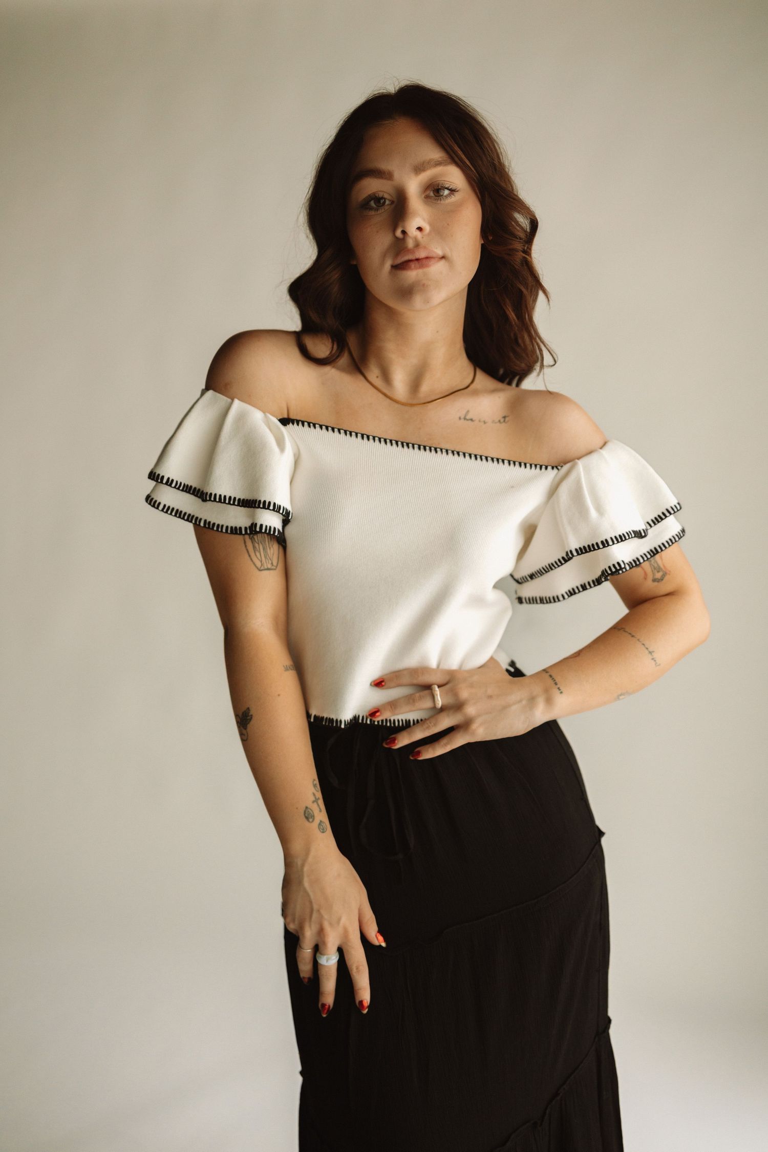 A person in an off-shoulder white top with black trim and black skirt poses against a neutral background.