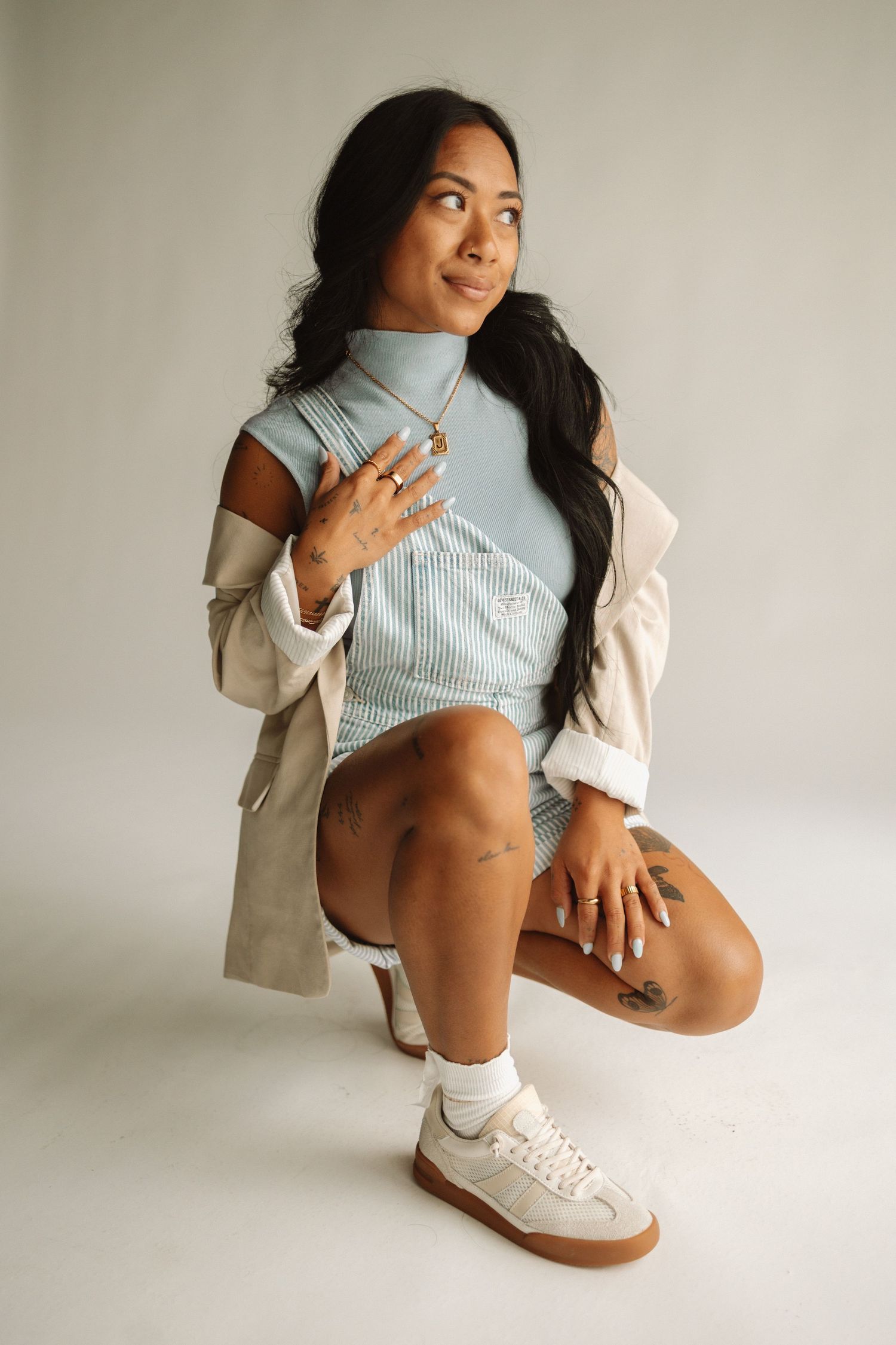 A person in a light blue shirt and beige blazer sits against a white wall and strikes model poses with white sneakers.