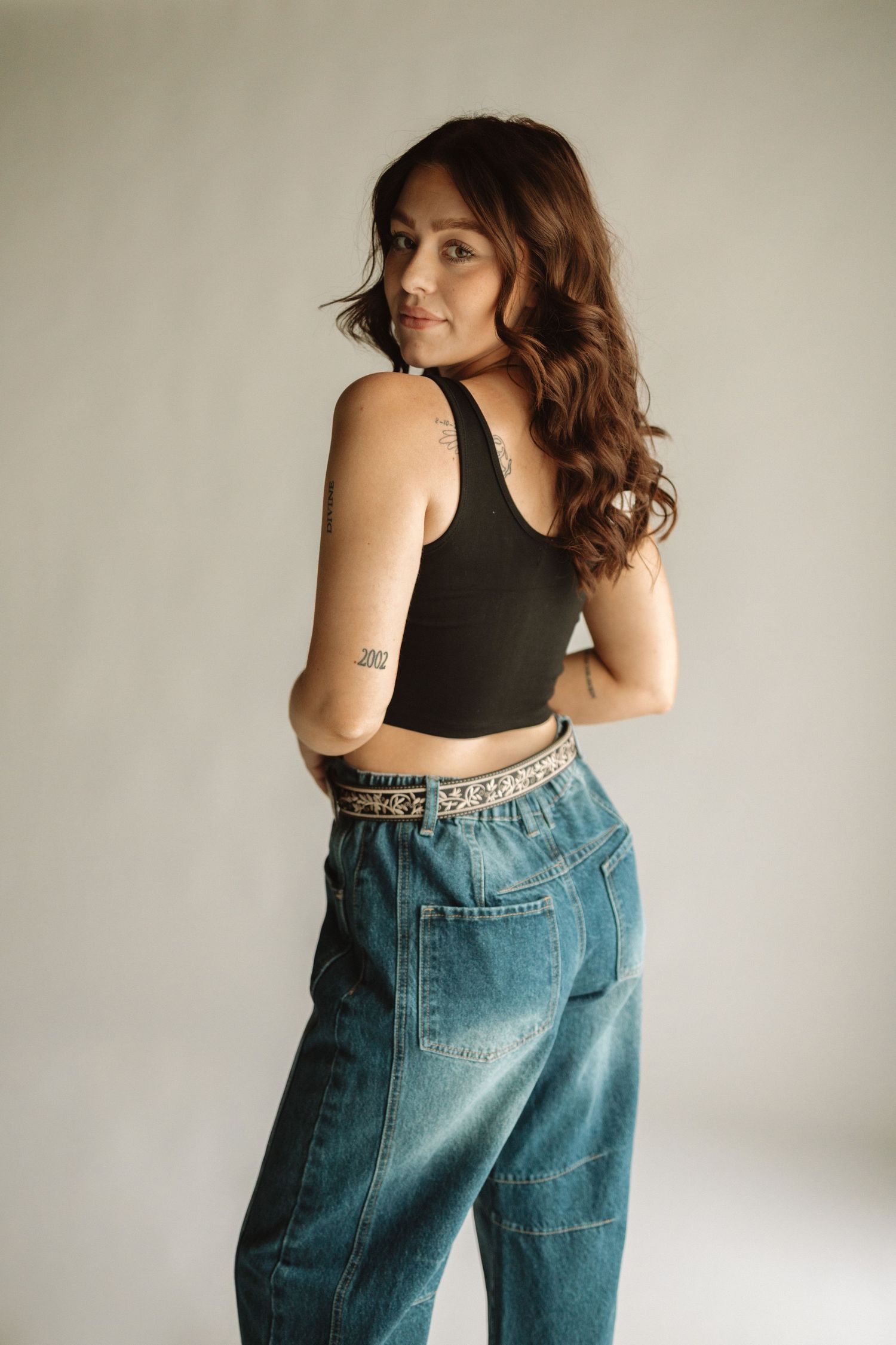 Someone in a black sleeveless top and baggy vintage blue jeans poses casually against a light background.