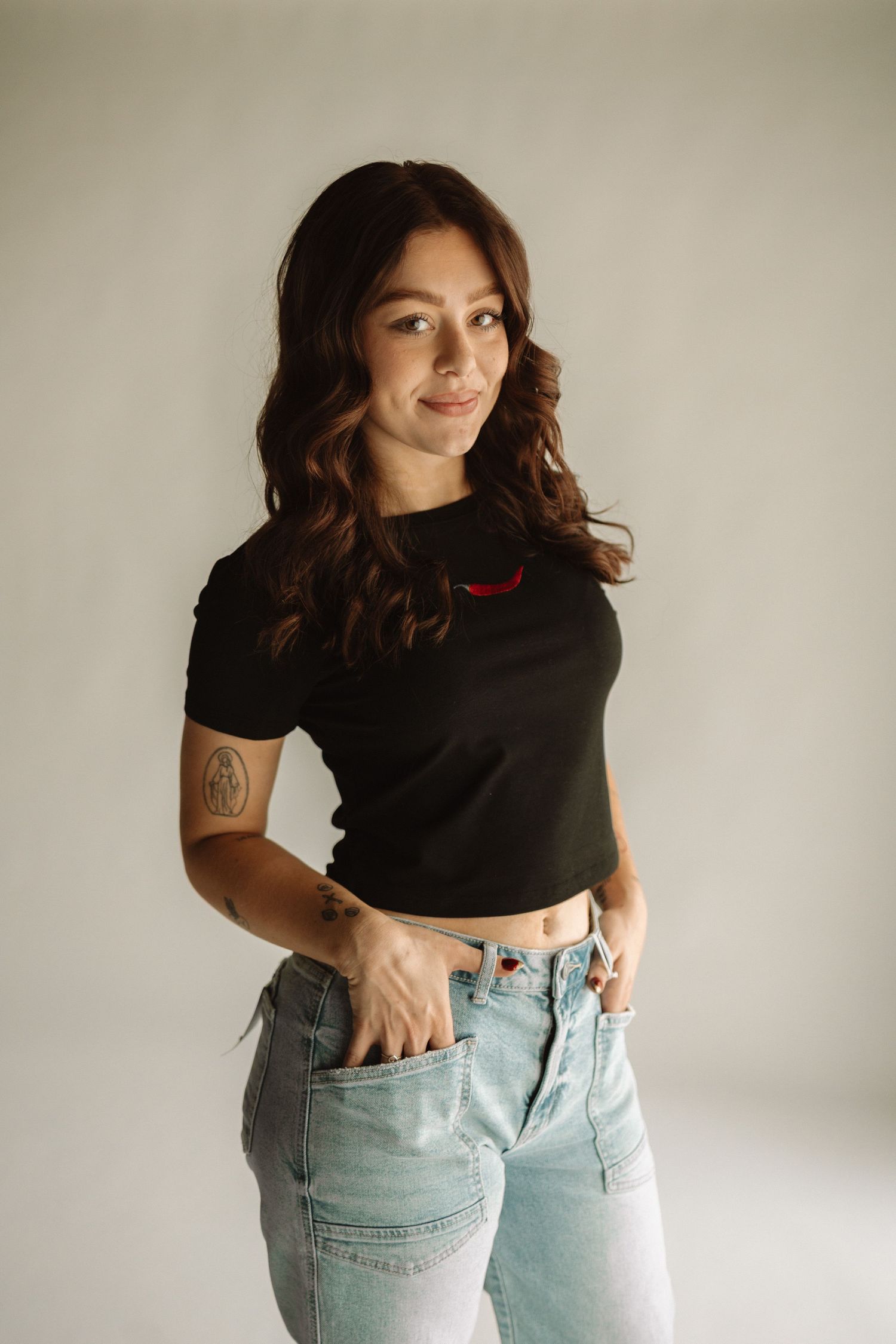 Studio portrait showing model in black crop top and light jeans with tattoo on arm posing against neutral background.
