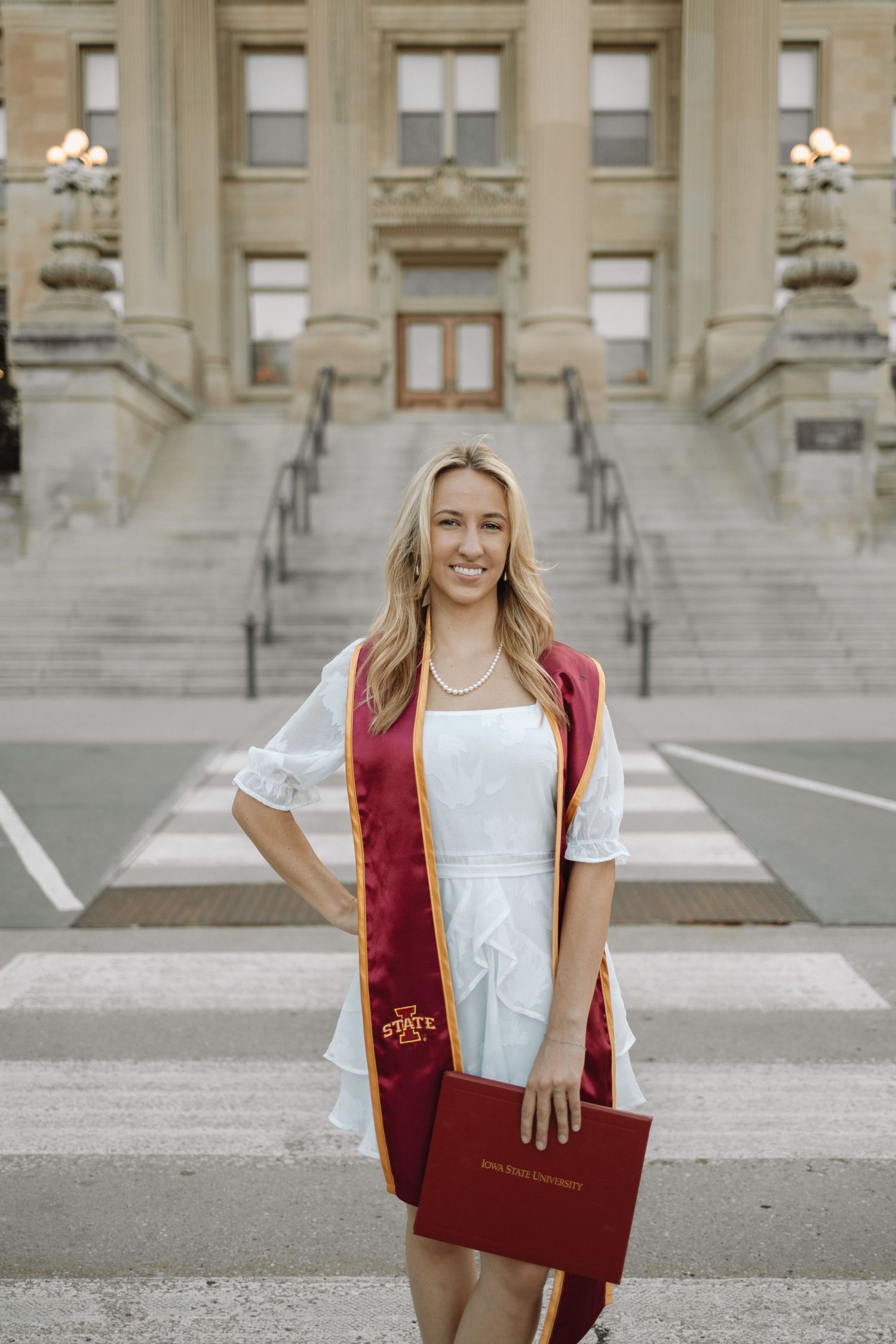 Graduate stands proudly on steps of historic building holding diploma in burgundy folder.