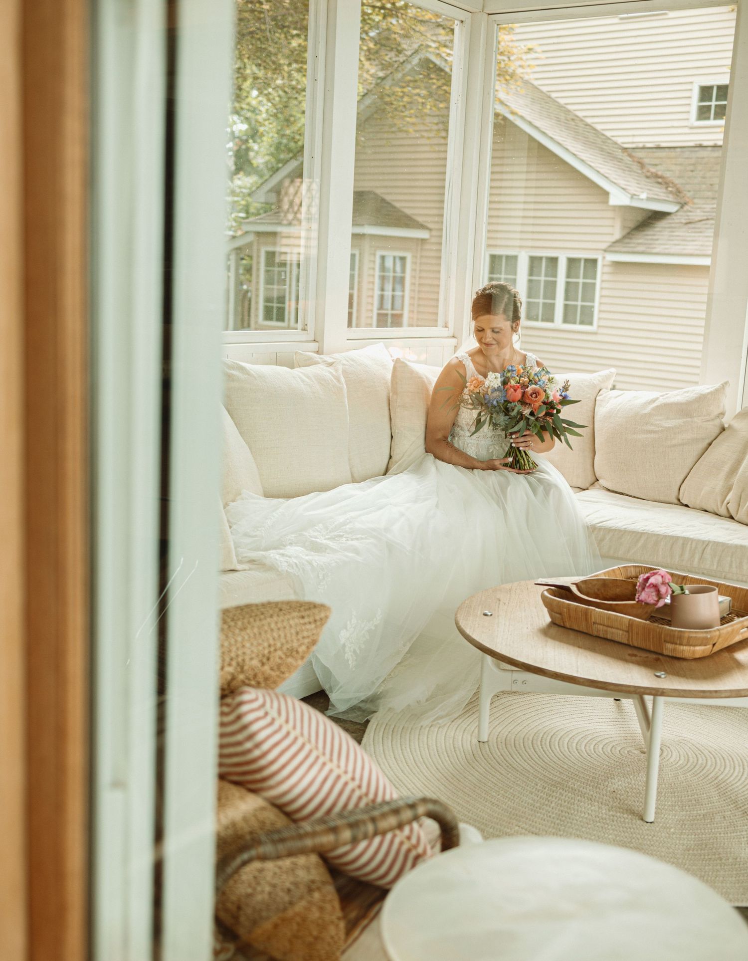 A bride in a flowing white gown sits in a sunlit sunroom with white furniture and holds a vibrant bouquet.