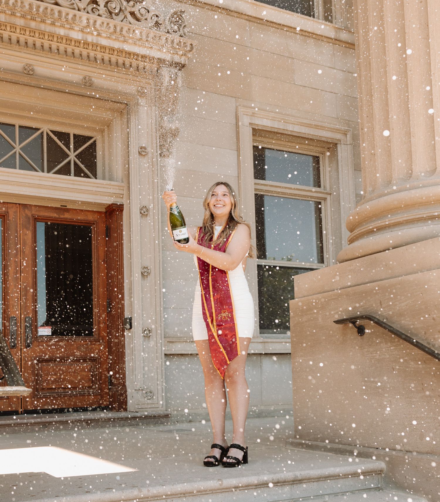 Someone in a red dress and white cardigan holding a champagne bottle on stone steps during a snowy celebration.