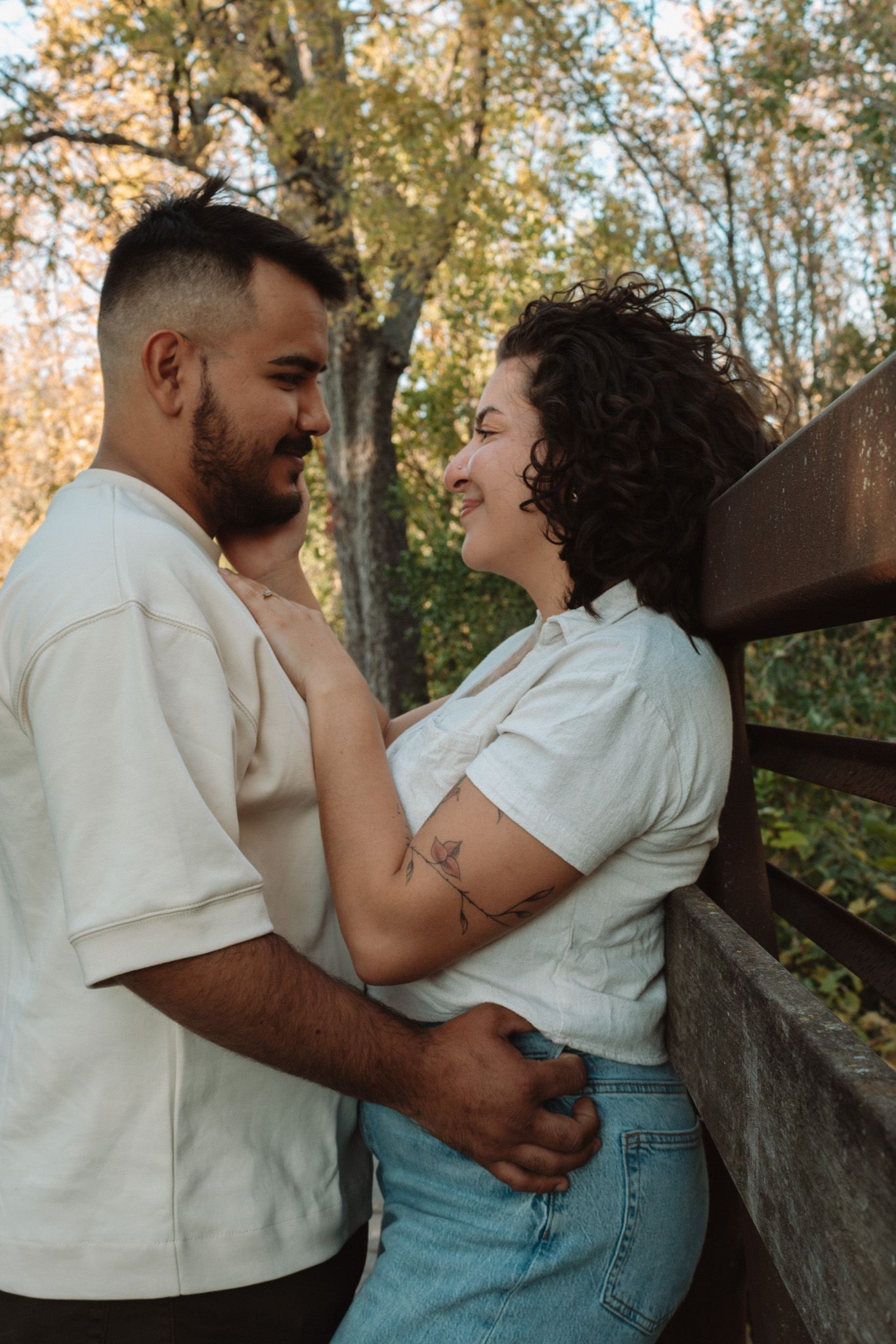 Couple in matching white tops sharing a tender embrace on a rustic wooden bridge with autumn foliage in background.