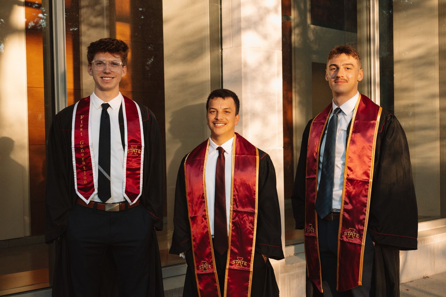 Three graduates in dark suits and burgundy stoles standing together in warm evening sunlight.