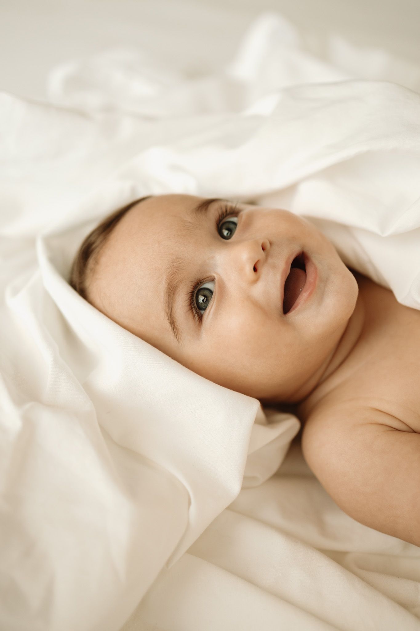 A baby lying on white bedding with wide eyes and an open mouth expression.