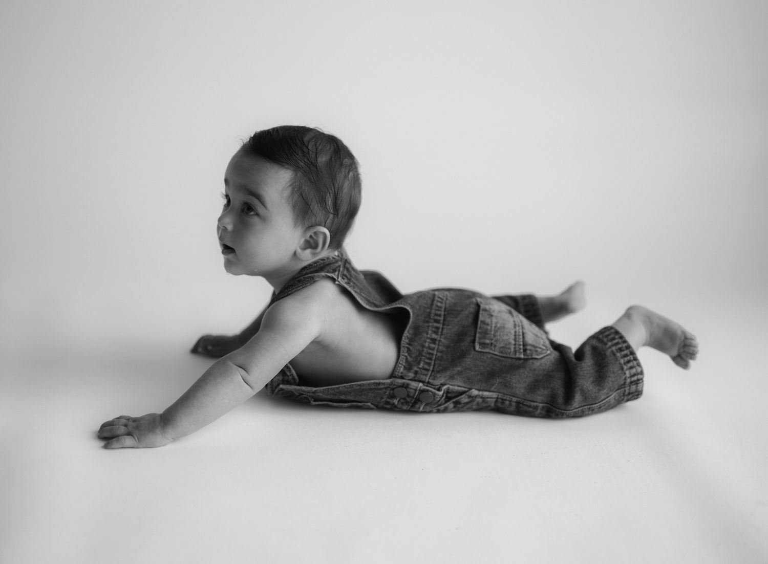 Black and white portrait of a baby lying on their stomach in denim overalls.