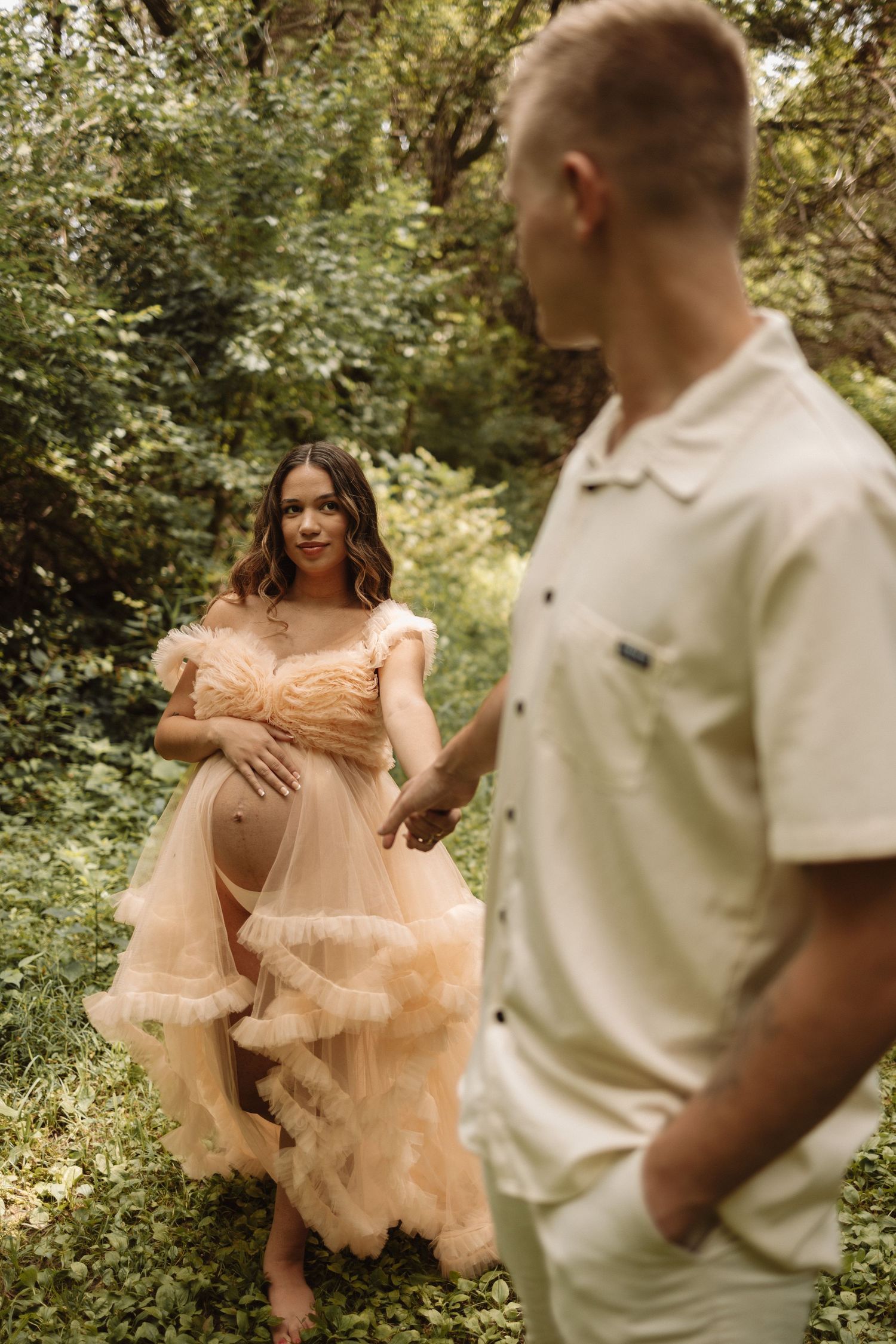 Expecting couple poses outdoors in a natural setting with person in peach gown showing baby bump among greenery.
