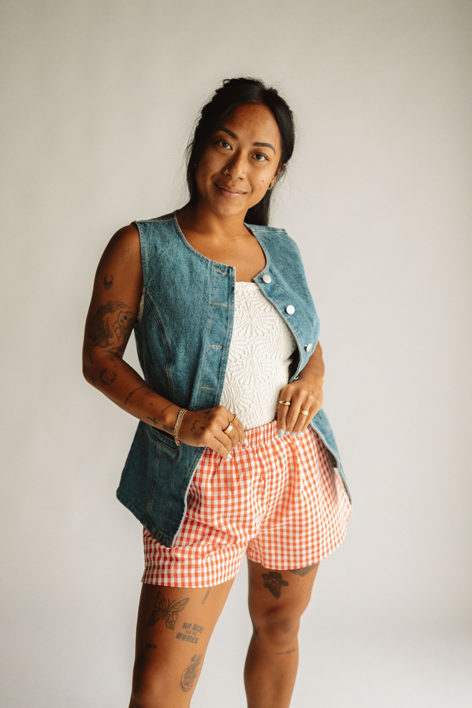 Person wearing denim vest, white lace top and orange gingham shorts poses against white background showing tattoos.