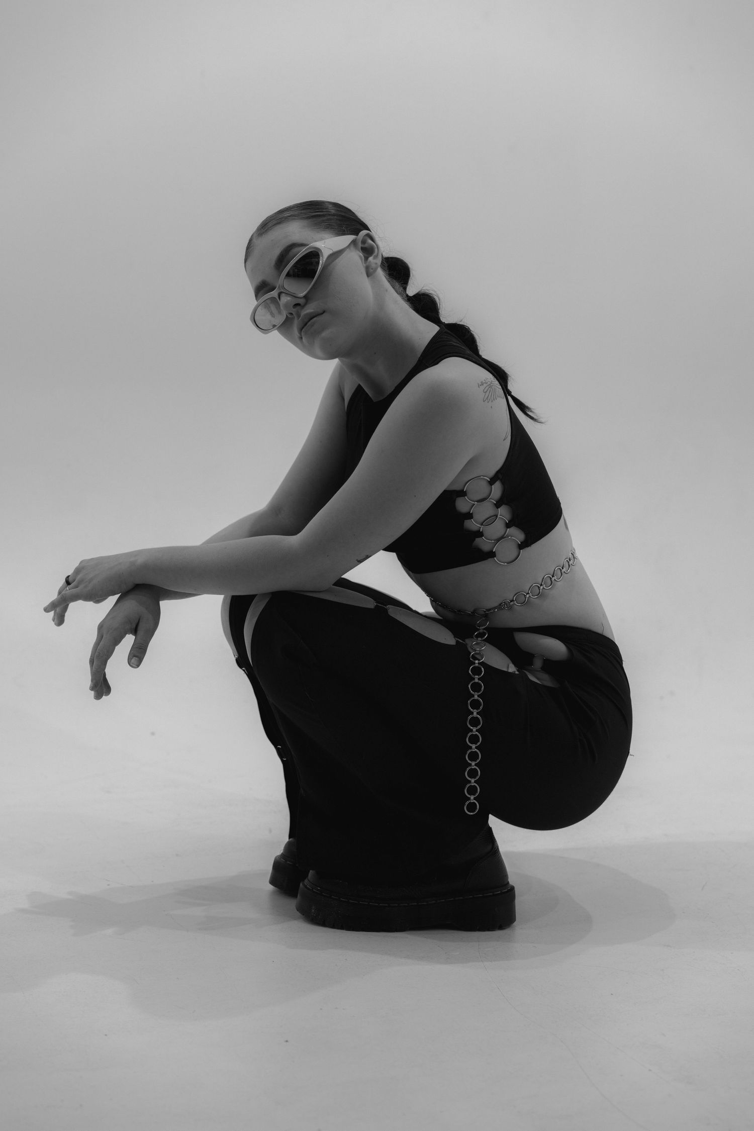 Black and white fashion portrait of person in sunglasses and crop top crouching in artistic pose against light backdrop.