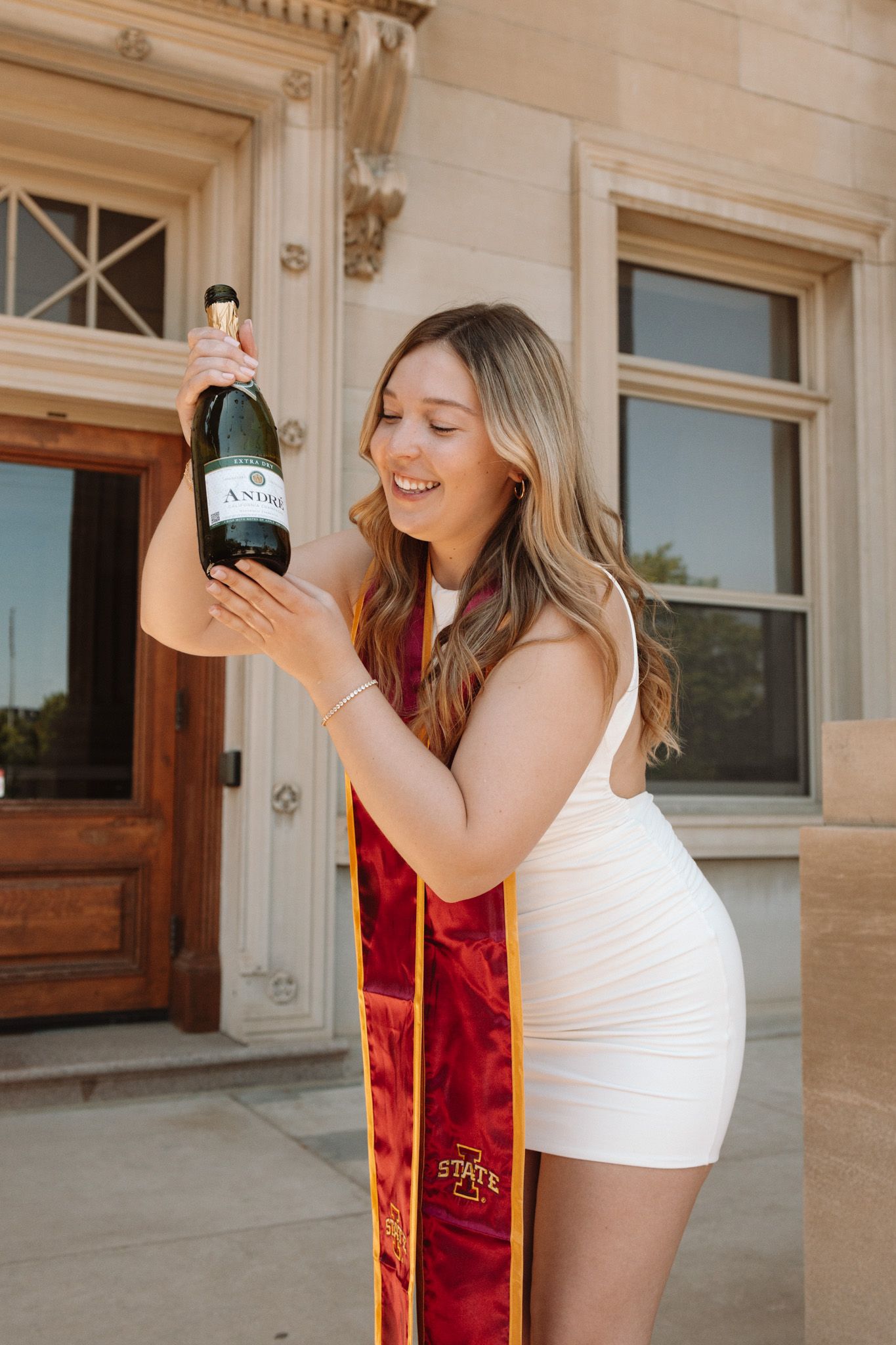 Graduate in white dress and red graduation stole celebrates with champagne bottle outside building.
