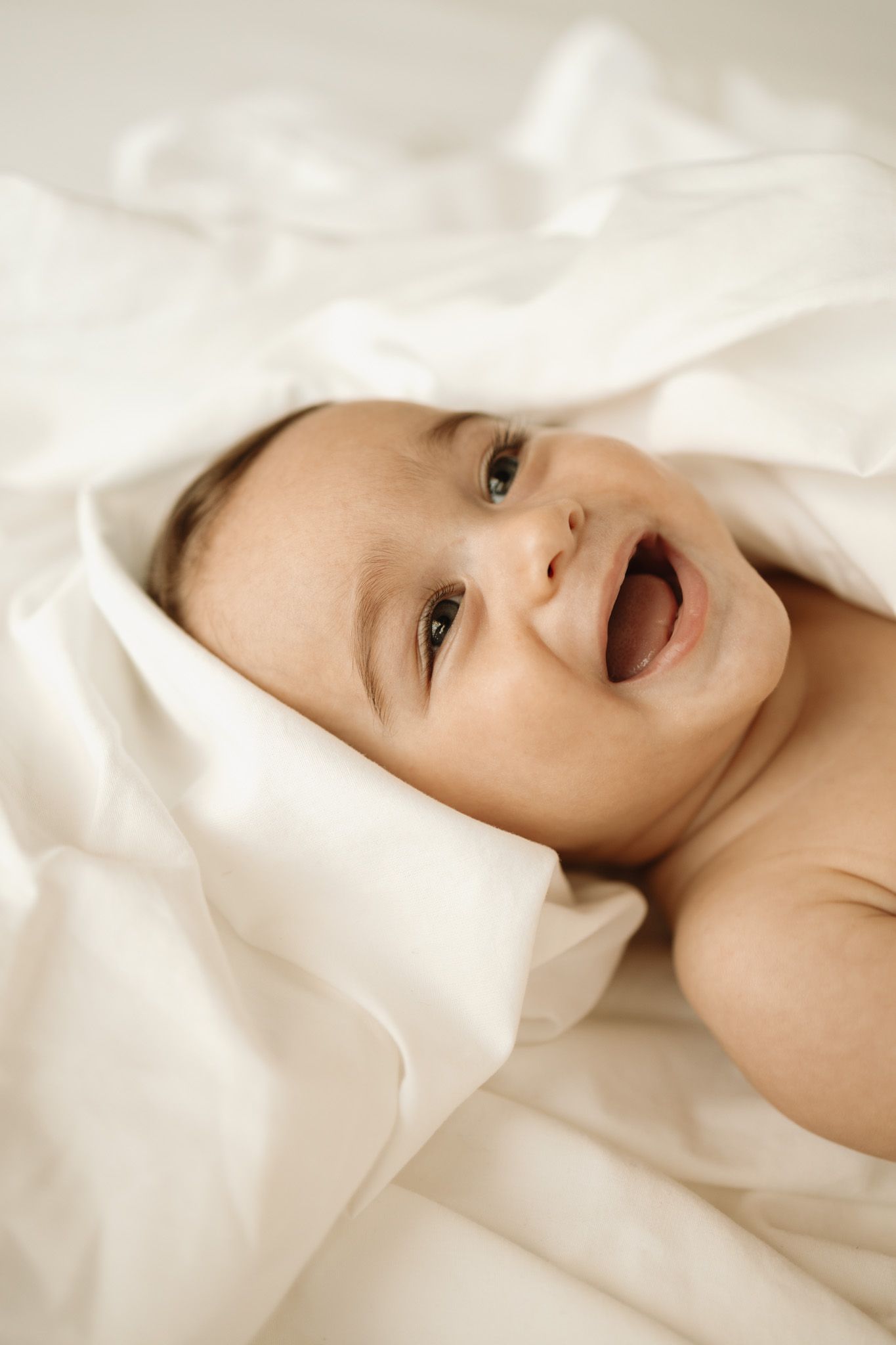 Happy baby lying on soft white bedding and blankets with a joyful expression and open smile.