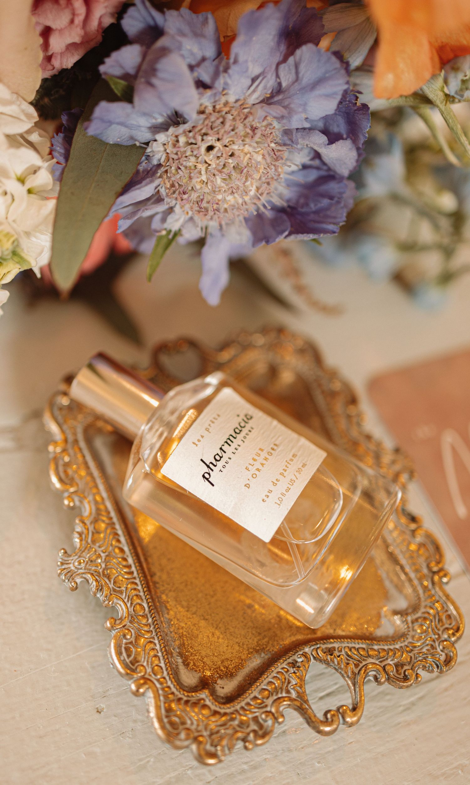 Elegant perfume bottle on ornate gold mirrored tray with delicate floral arrangements in the background.