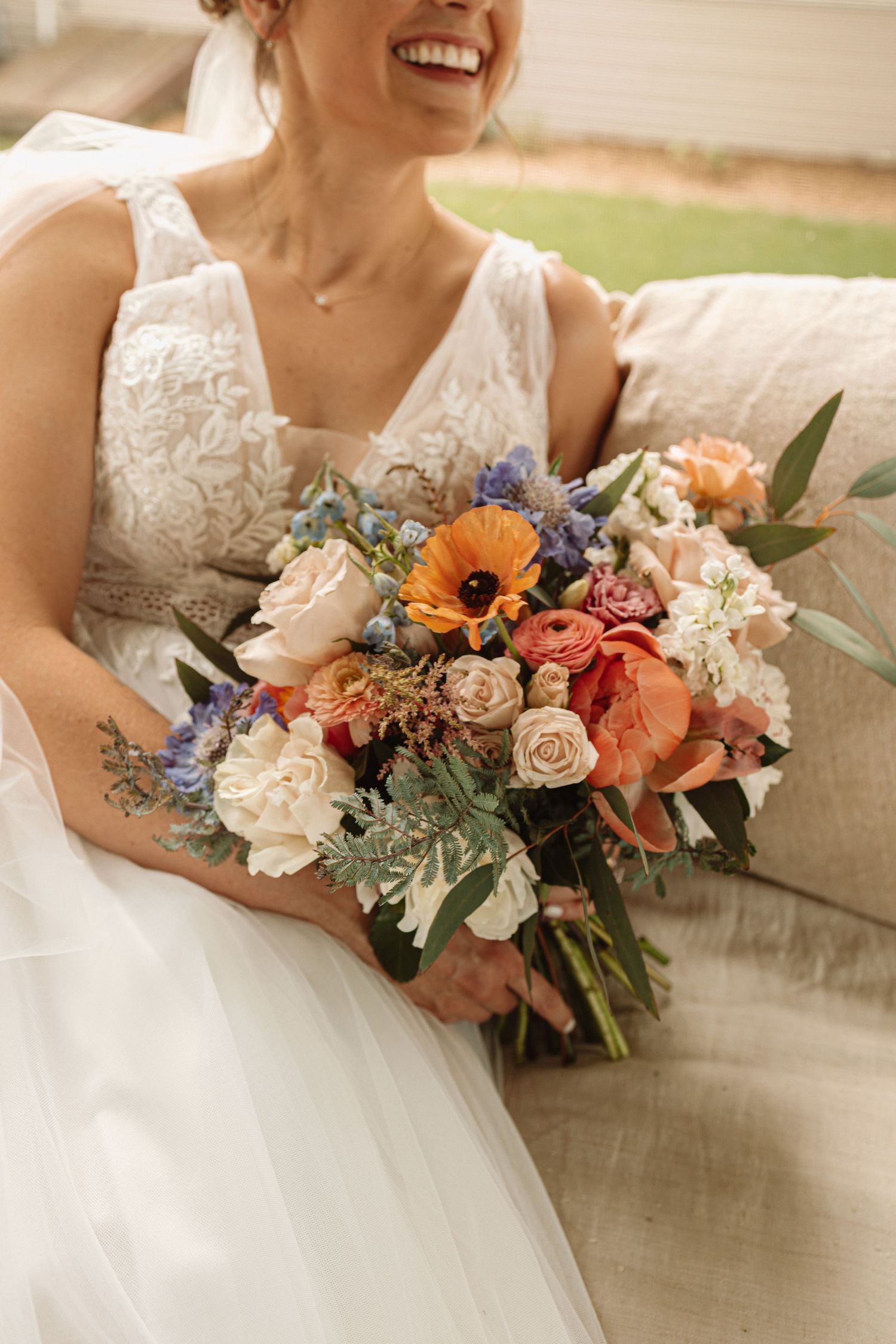 Wedding bouquet featuring peach, coral and blue blooms with greenery held against a white lace dress.