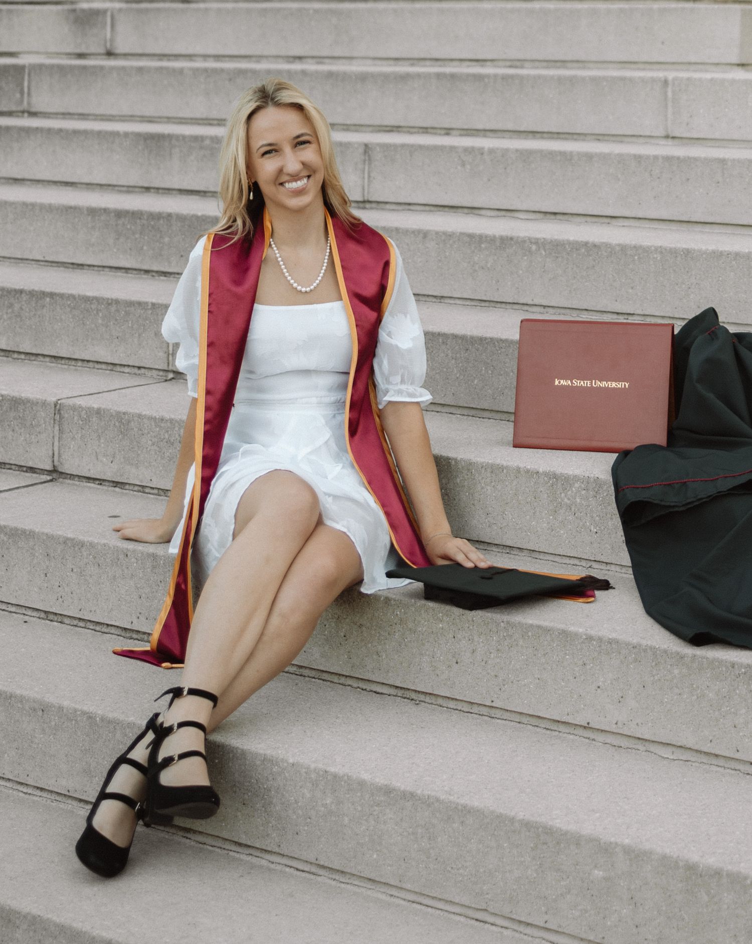 A person in a white dress and red vest sits casually on concrete steps with a bag nearby.