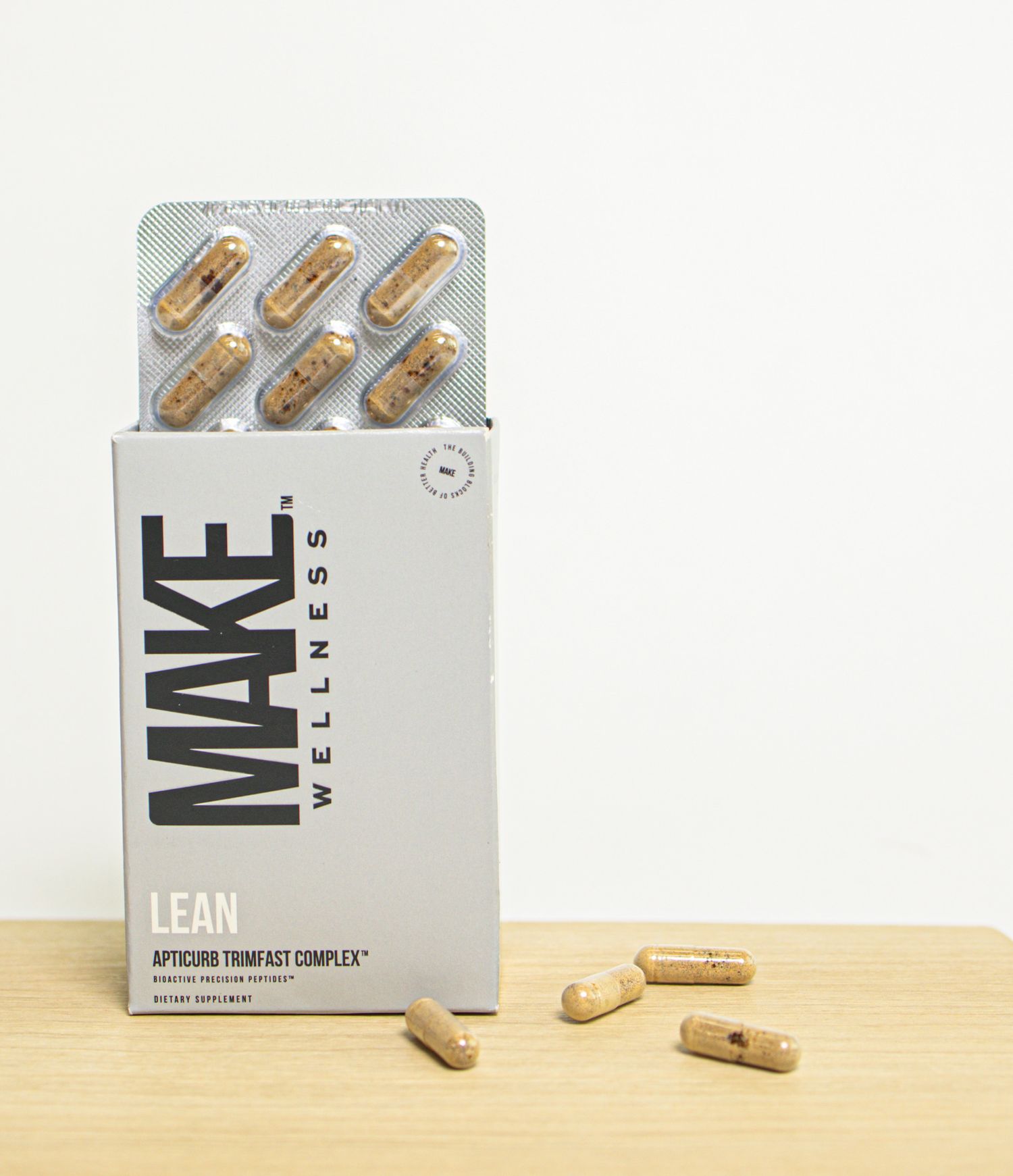 A bottle of MAKE Wellness supplements with beige capsules displayed on a wooden surface against a white background.