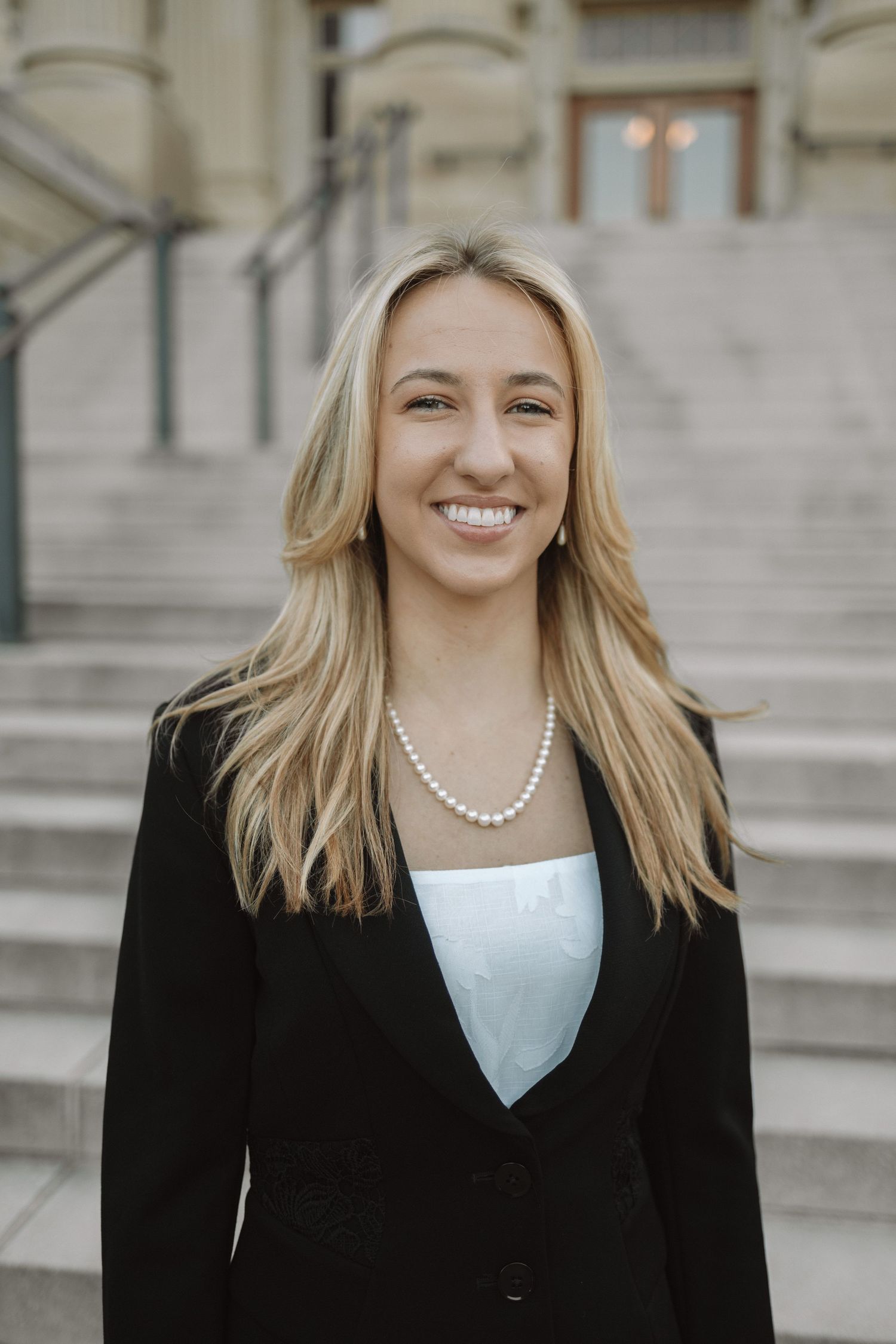 Professional portrait of someone in a black blazer and pearl necklace standing on stone steps of a building.