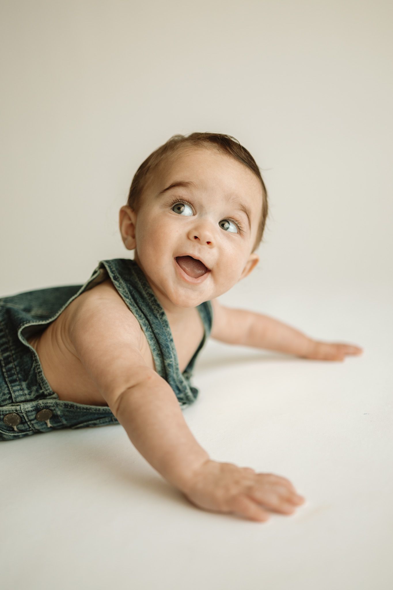 Adorable baby in denim overalls crawls on white background while looking up with a joyful expression.