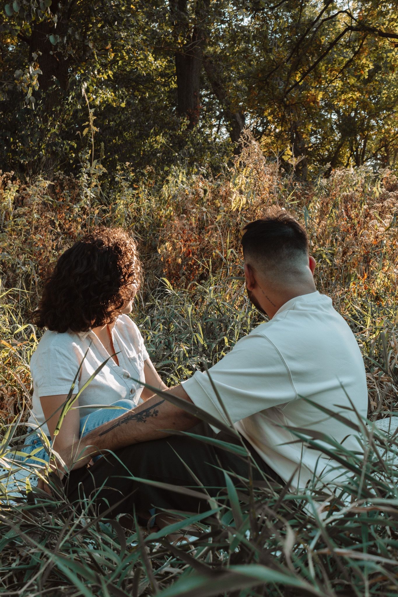 Two people sit together in tall grass and foliage during golden hour, sharing an intimate outdoor moment in nature.
