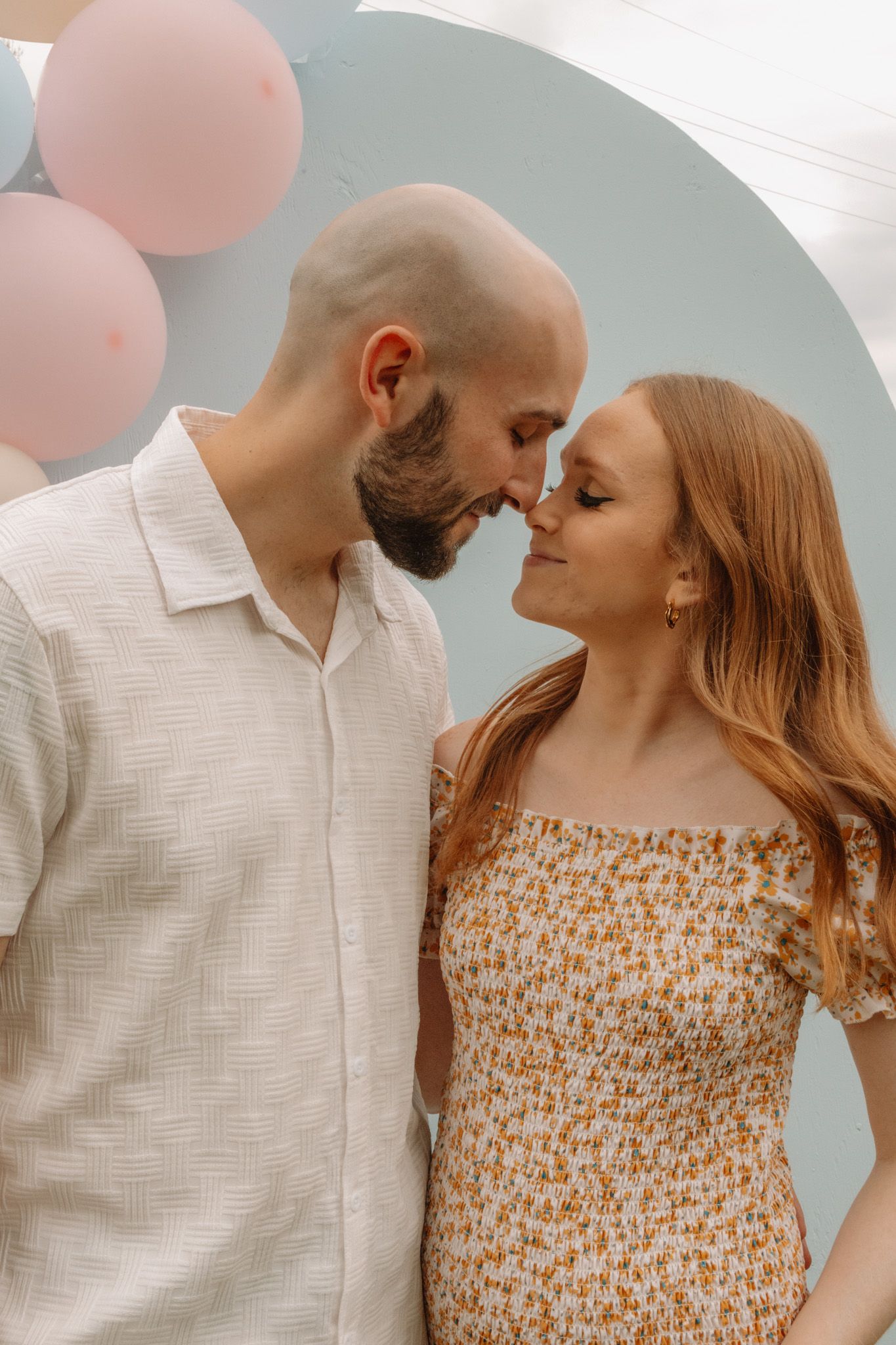 A romantic couple shares an intimate moment in front of a pastel backdrop with pink balloons.