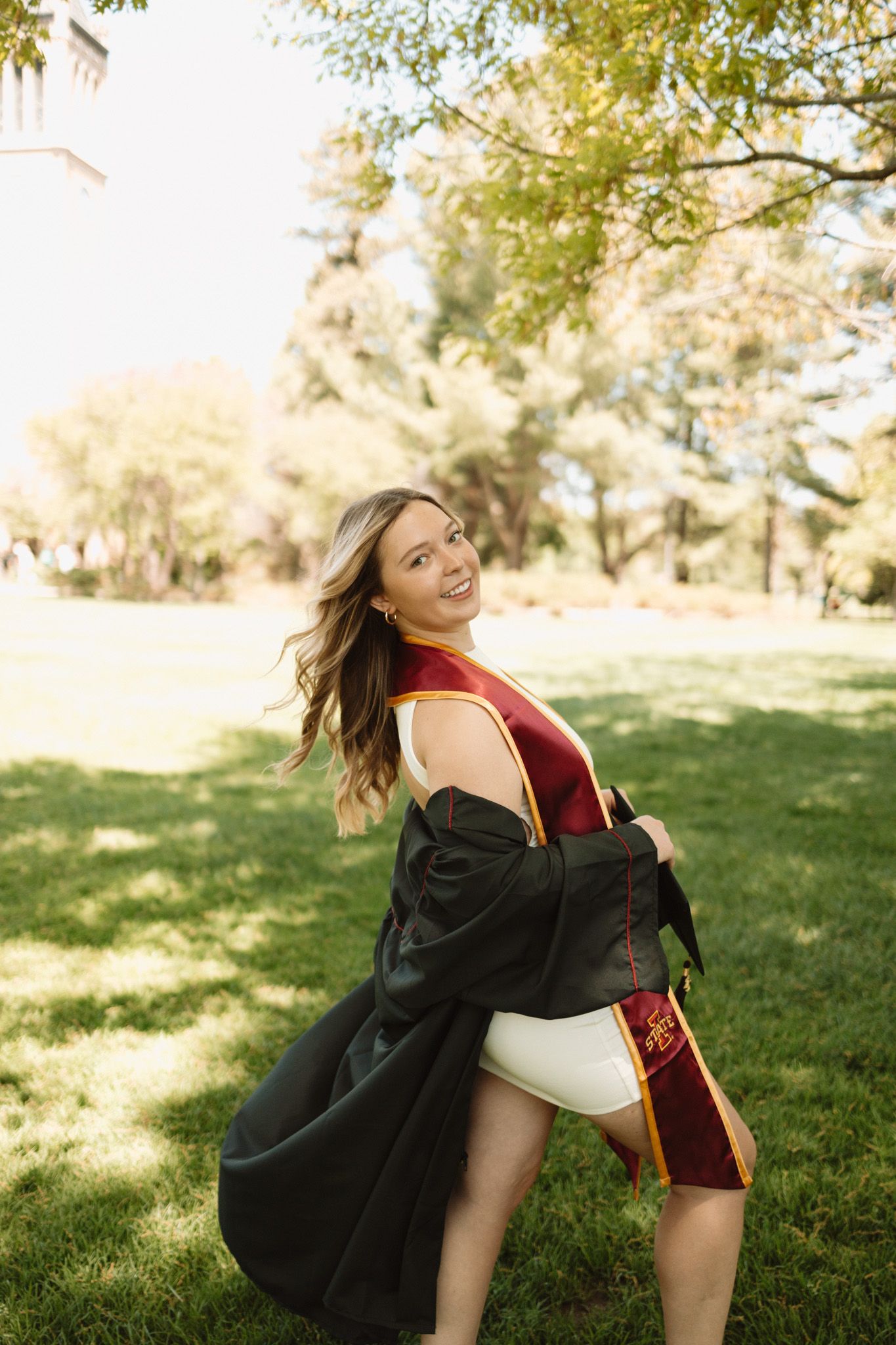 A graduate in a red gown and black robe twirls playfully in a sunny park setting during a graduation photoshoot.