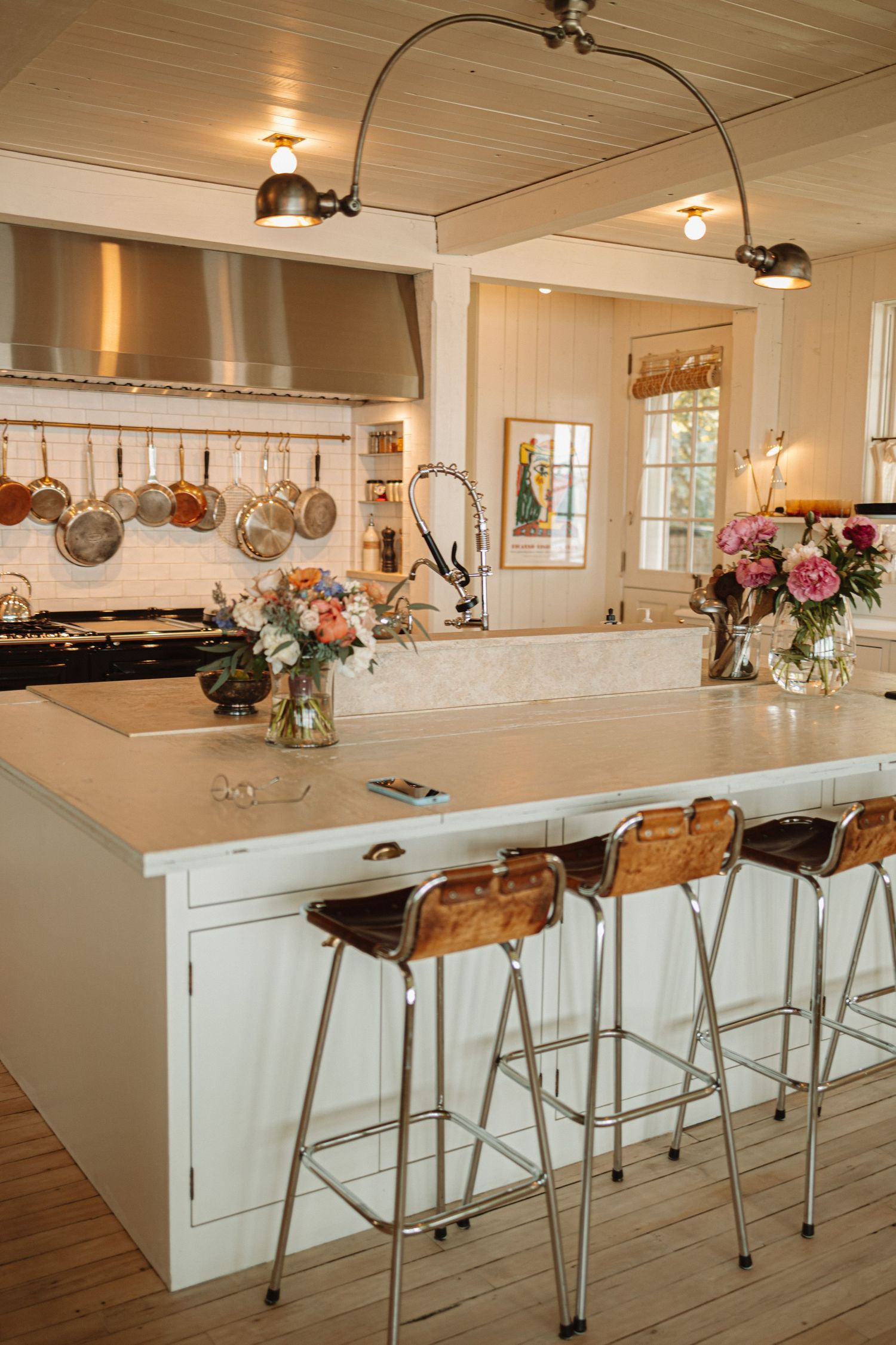 Modern white kitchen features a stylish bar with metallic stools and hanging copper cookware in a rustic-chic setting.