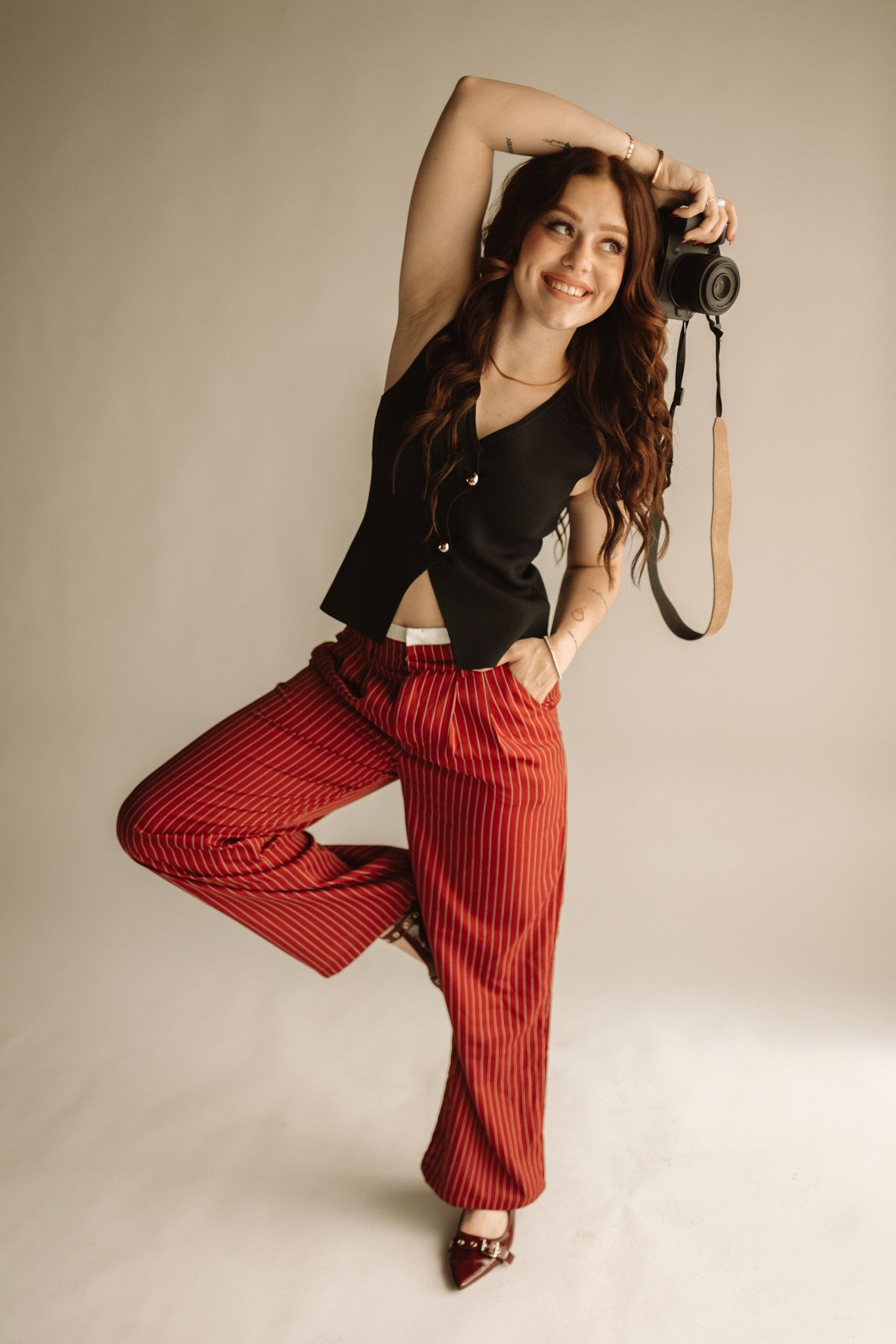 Fashionable photographer poses in black crop top and red striped pants while holding a vintage camera against beige backdrop.