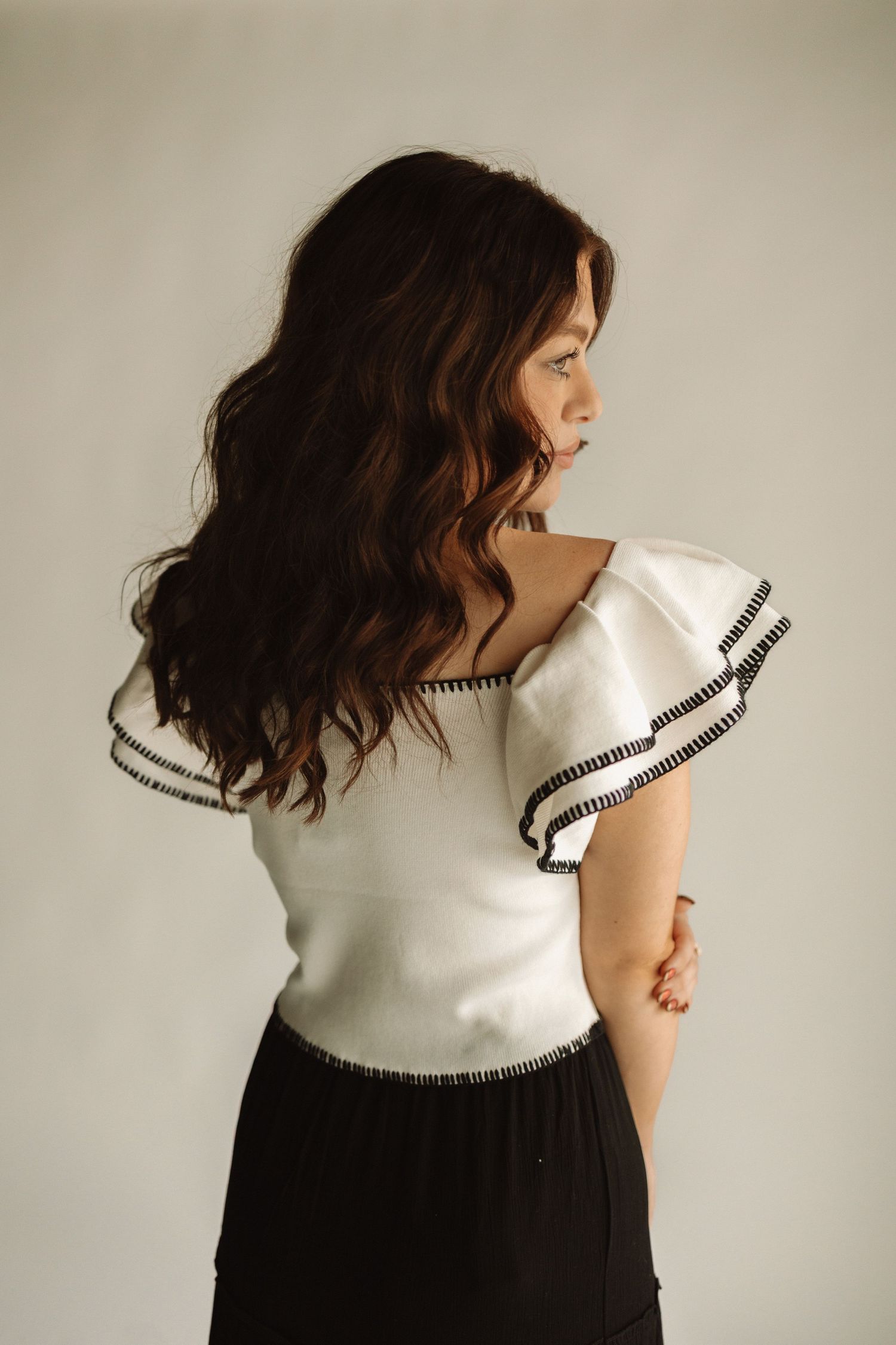 Woman with wavy brown hair wearing a white top and black skirt poses against a neutral background.