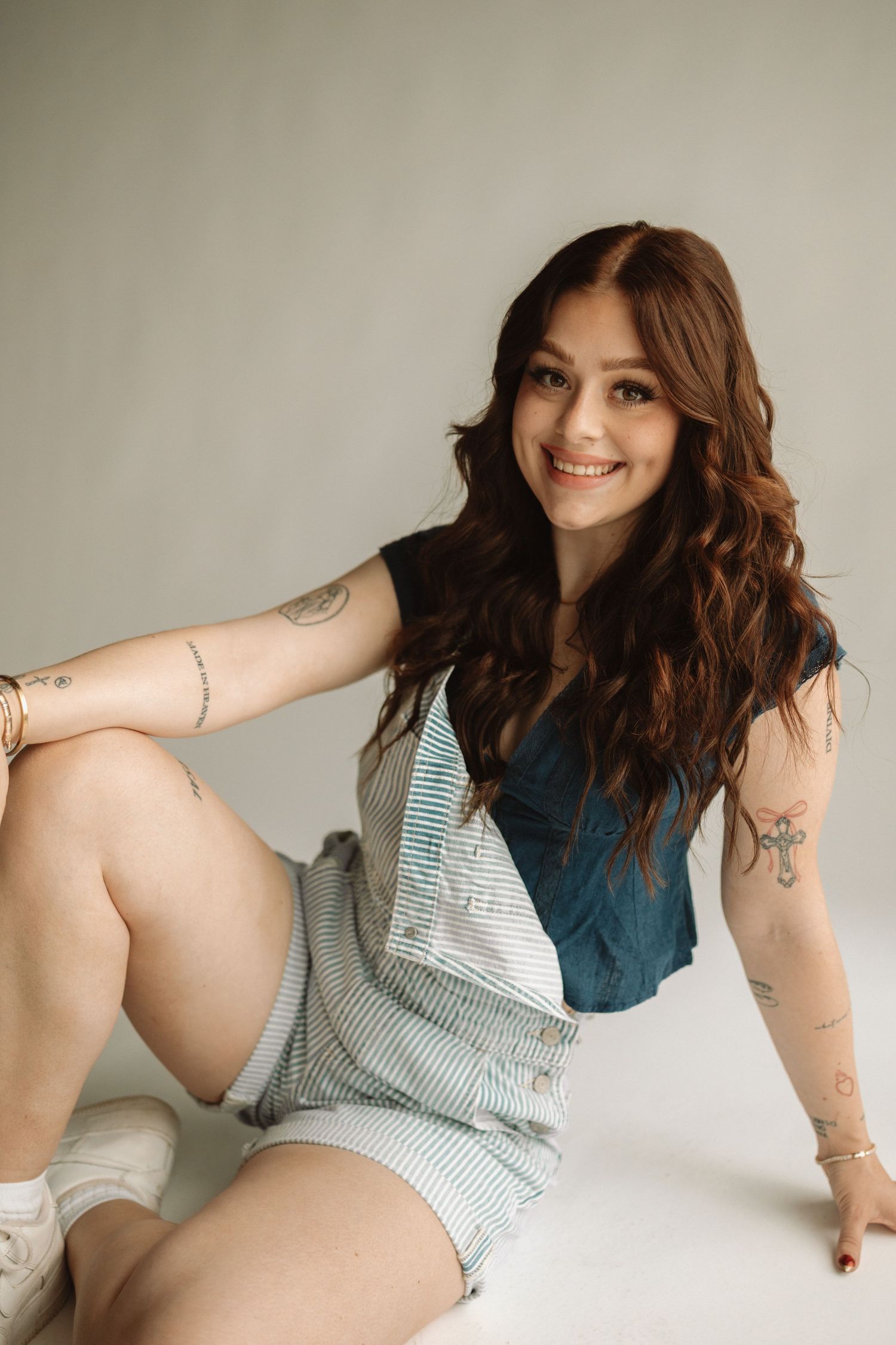 Woman in striped swimsuit poses on light background showing curves and tattoos in artistic boudoir series.