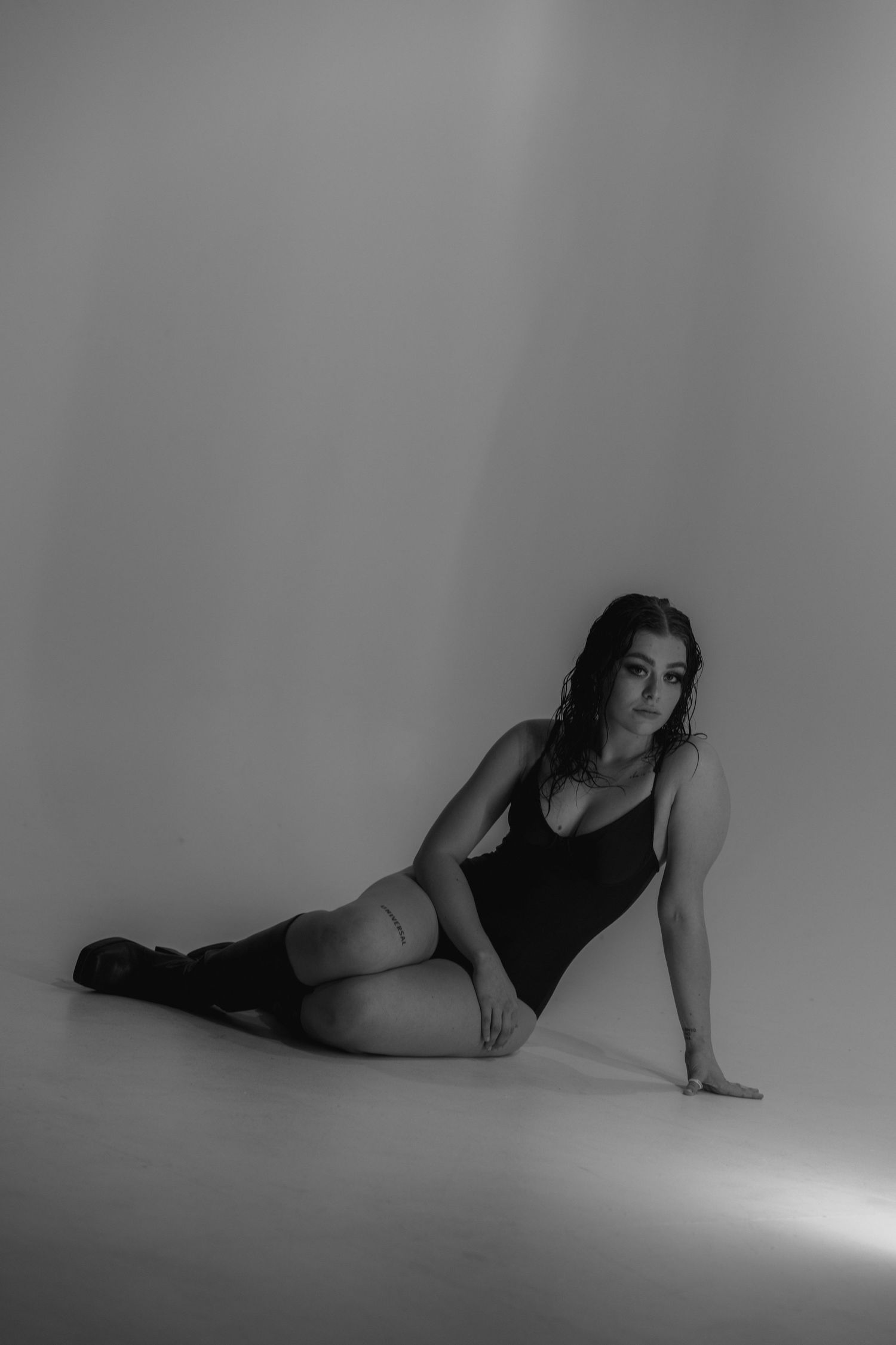 A black and white artistic studio portrait of a woman in a bodysuit posing on the floor.