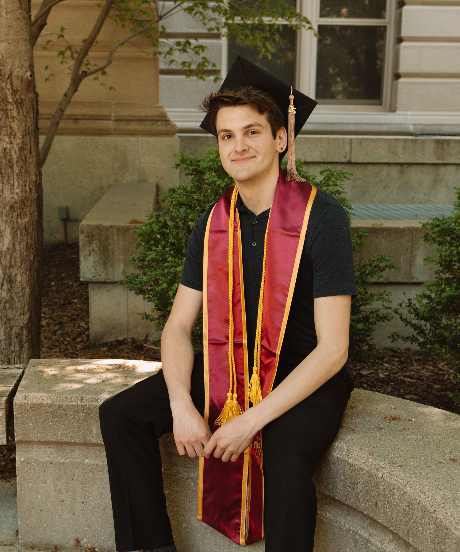 Graduate in black academic dress with maroon and gold stole poses on stone steps for portrait series.