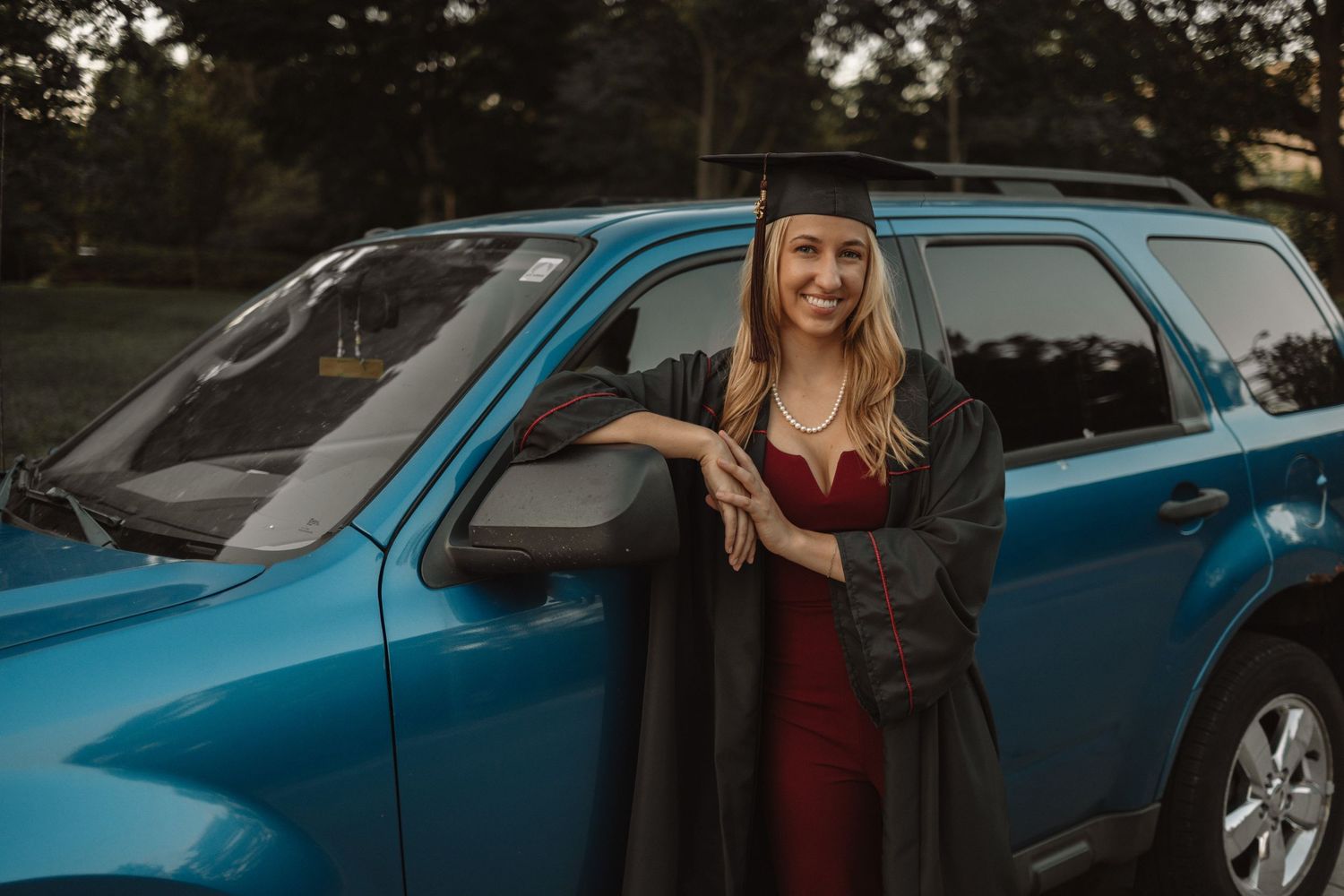 A metallic blue car is parked outdoors with someone posing beside it in burgundy clothing.