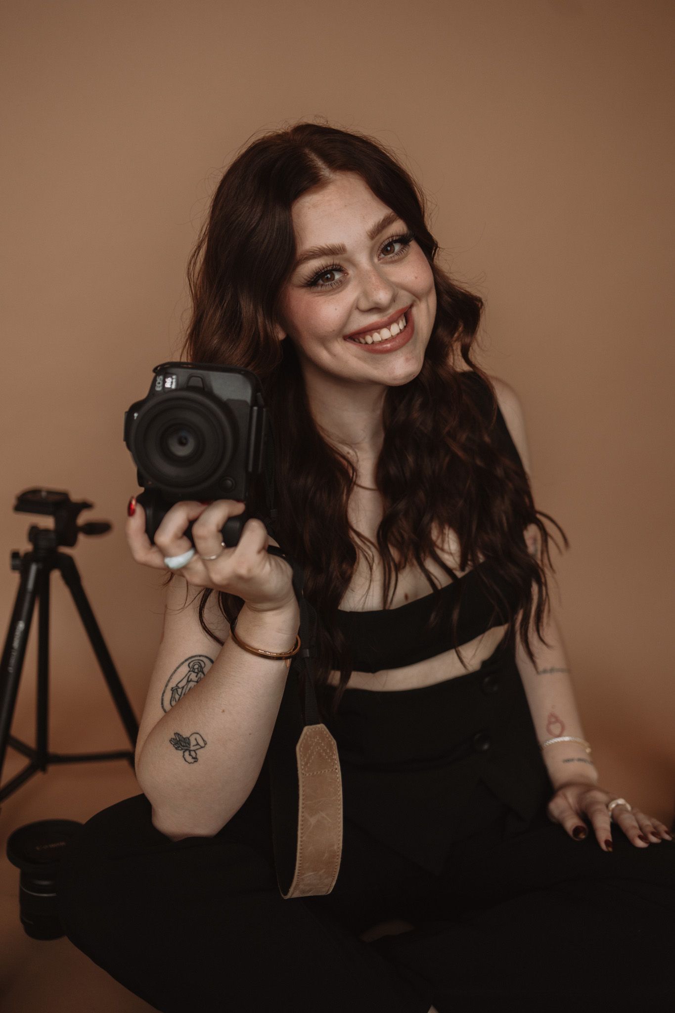 Portrait of a photographer with dark hair holding a vintage camera while sitting against warm brown backdrop.