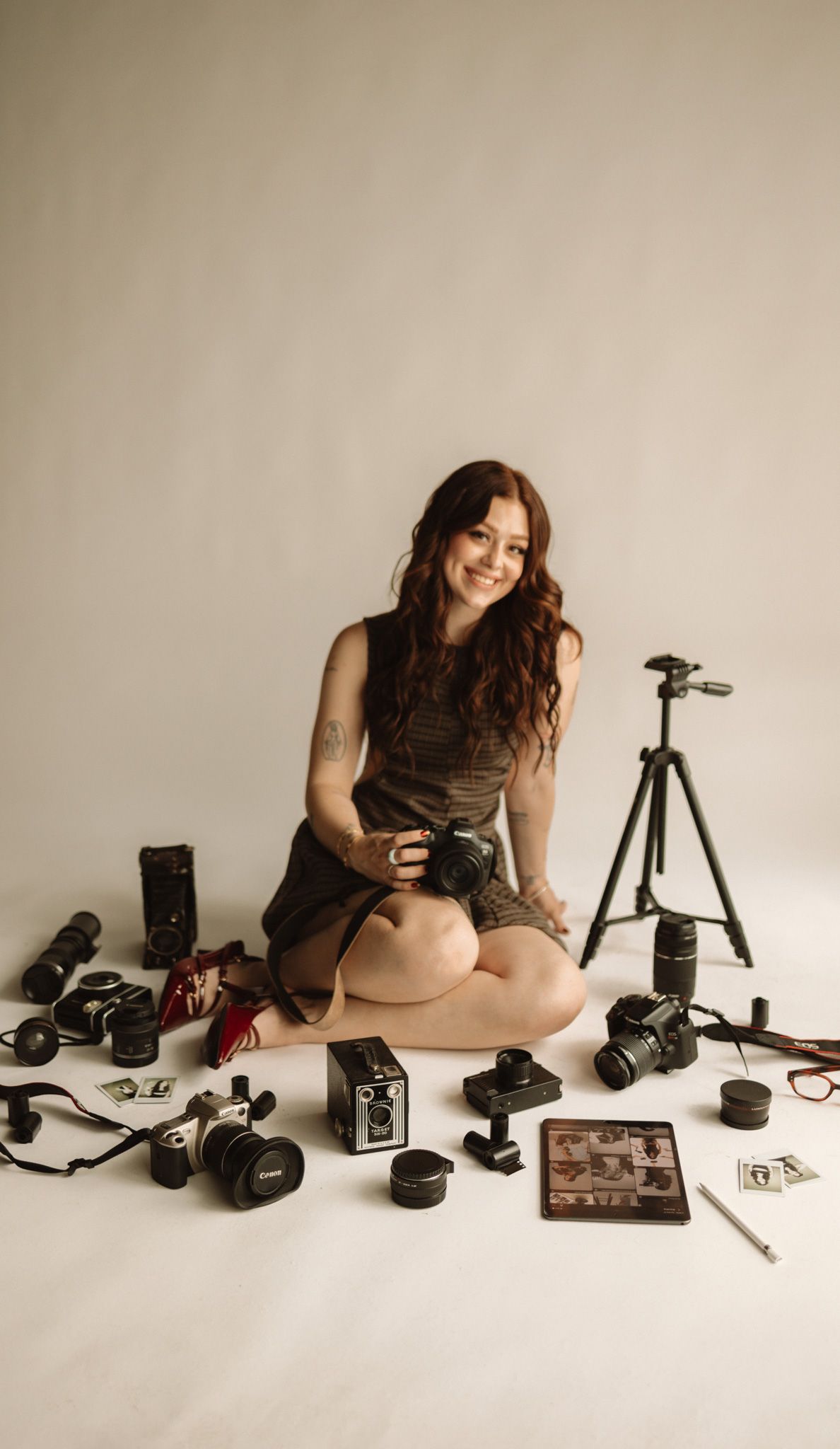 Photographer sits surrounded by professional camera equipment including lenses, tripod and digital tablet on white backdrop.
