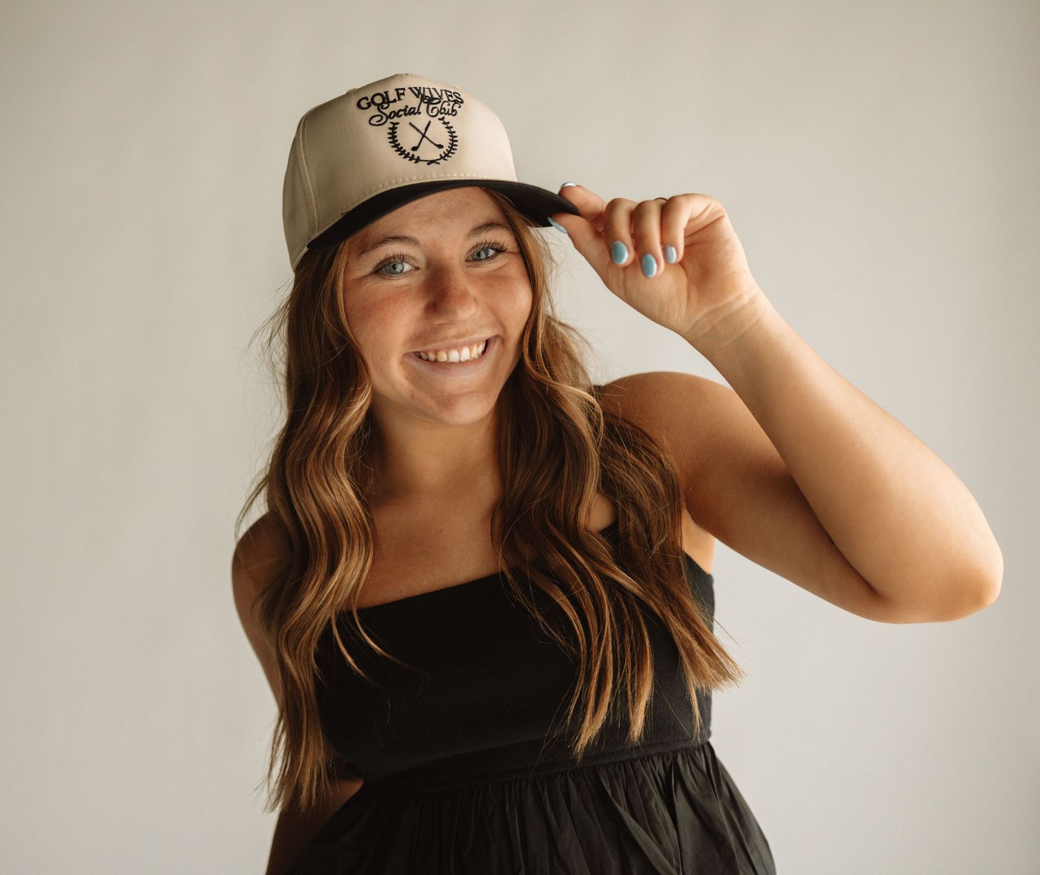 Cheerful person wearing beige baseball cap with wavy brown hair and black top against white background.