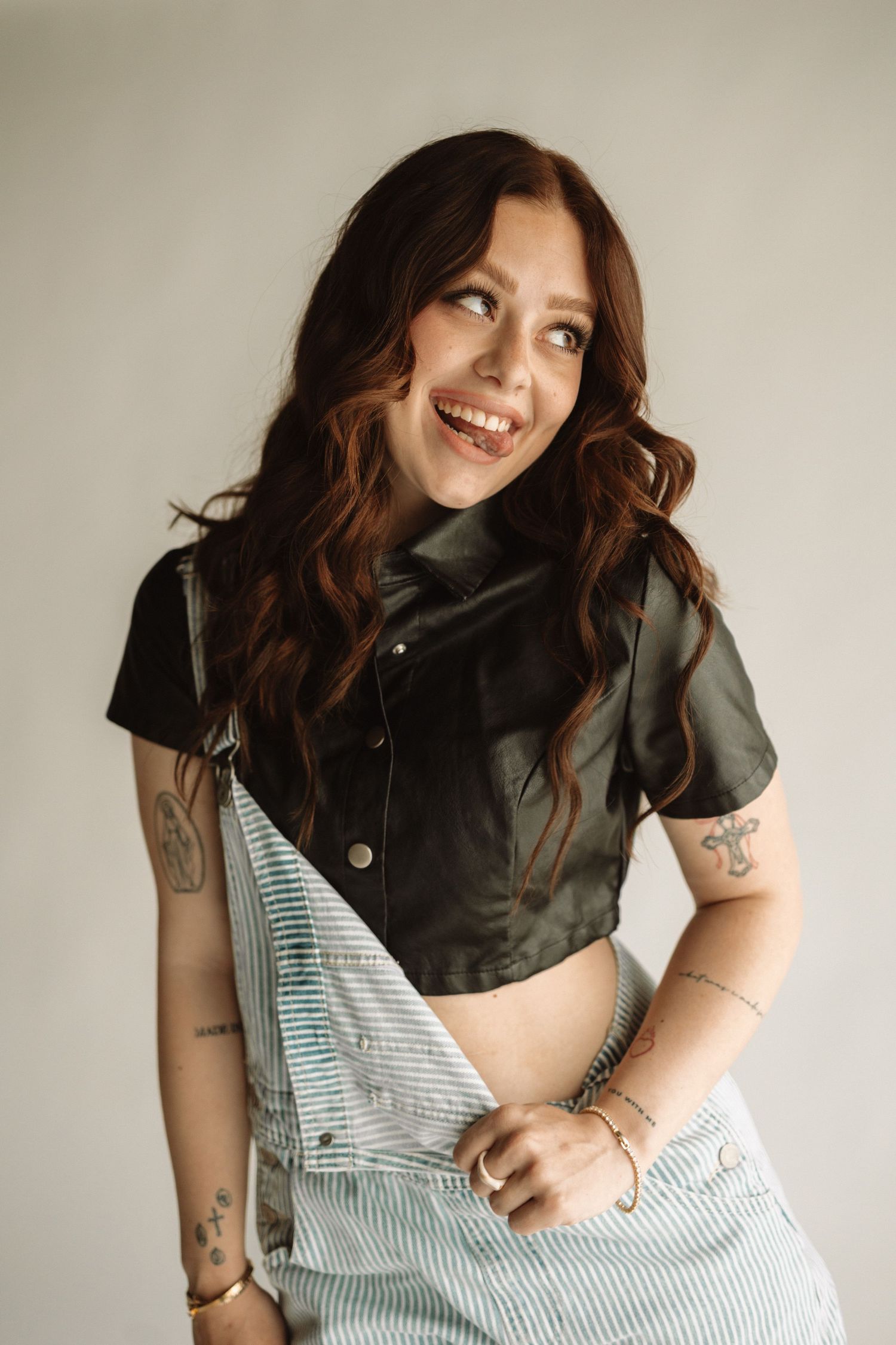 Cheerful woman wearing striped overalls and black shirt poses against white wall with brown wavy hair and tattoos.