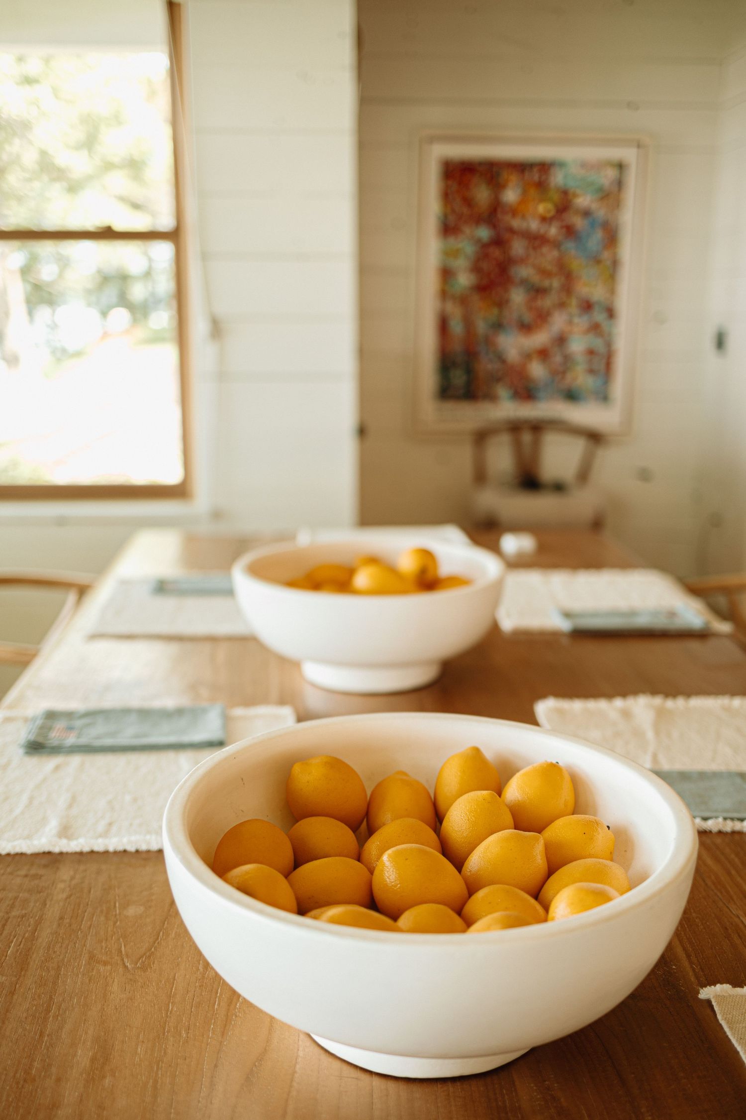 Fresh tangerines arranged in a white ceramic bowl on a wooden table with framed artwork in the bright background.
