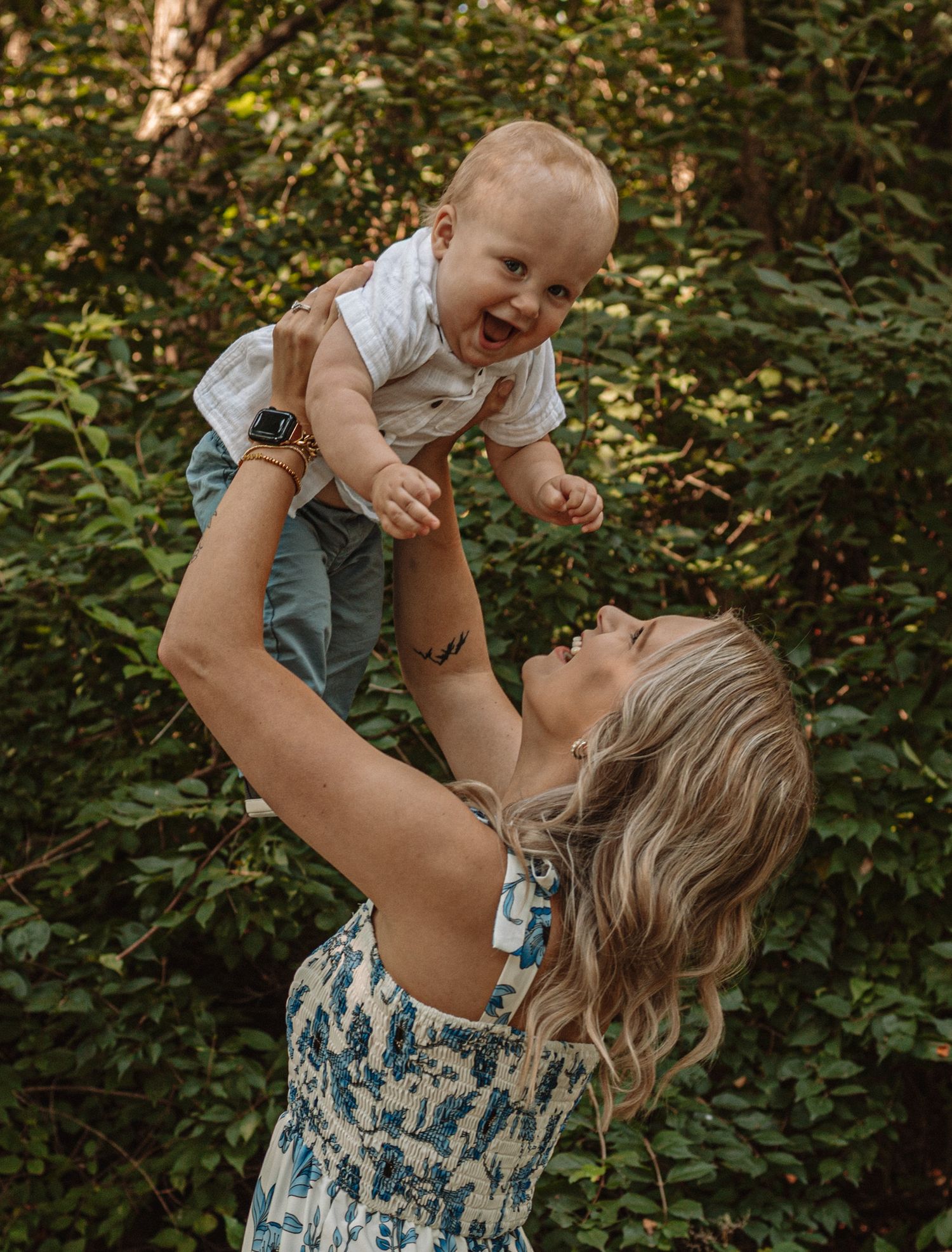 A joyful outdoor moment captured in a forest setting with lifted baby wearing light colored clothing.