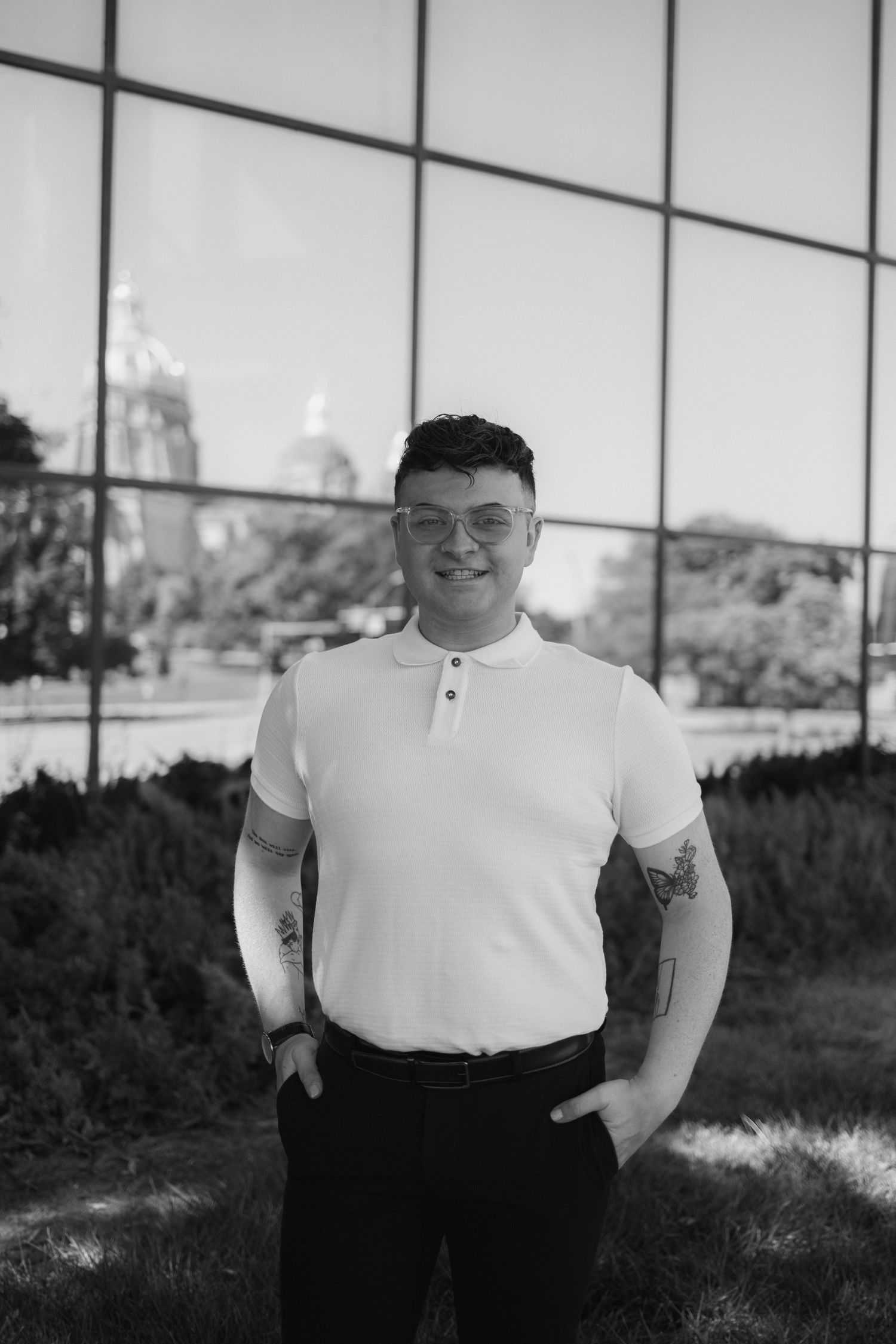 A person in a white t-shirt stands outdoors against a glass building facade in black and white photography.