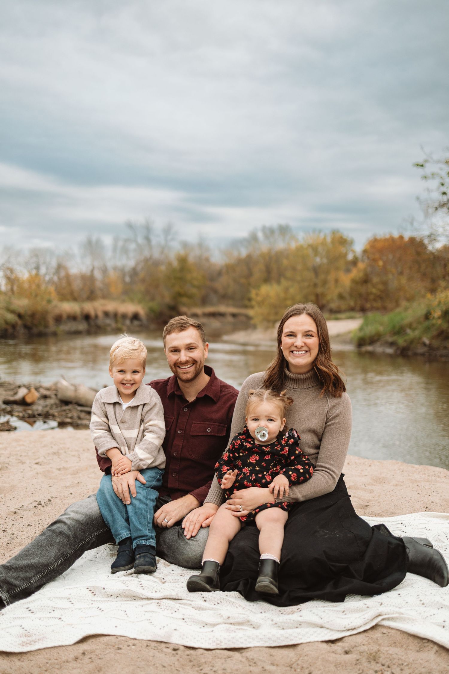 Family sits together on white blanket by river bank during autumn outdoor photo session.