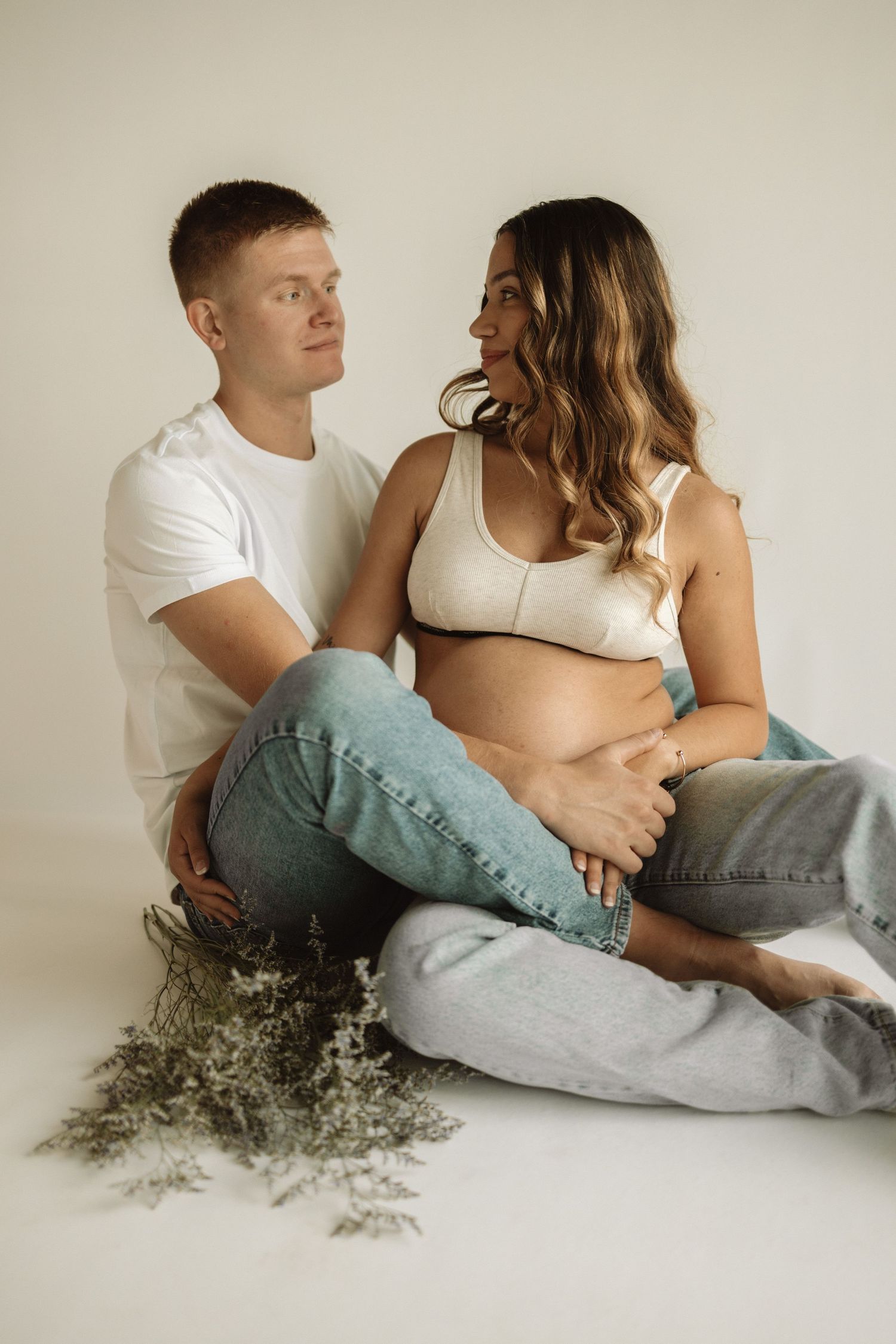 A couple in casual white and denim outfits reclines together on a white background with delicate greenery.