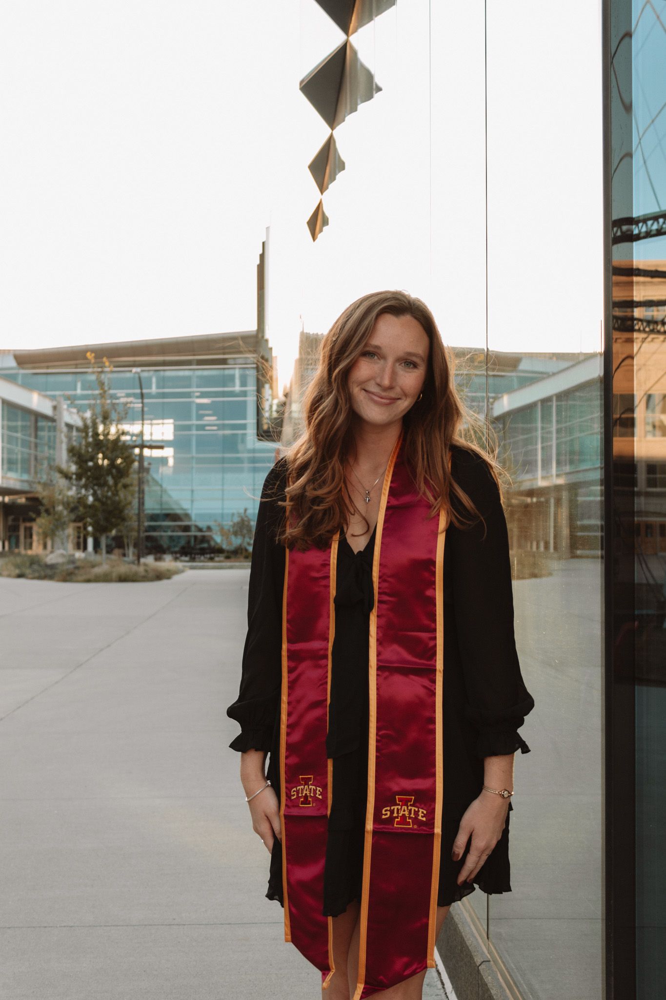 Graduate wearing red and gold graduation stole and black dress standing outside modern building with glass walls.