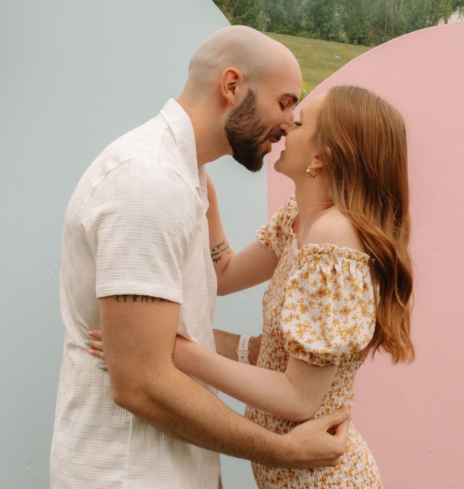 A romantic couple embraces outdoors, wearing summer attire with a floral dress and white shirt against a natural background.