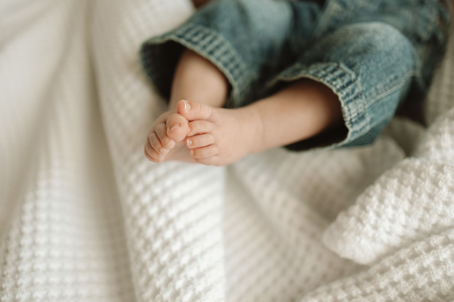 A small baby's feet wrapped in soft knit denim booties against a white crocheted blanket background.