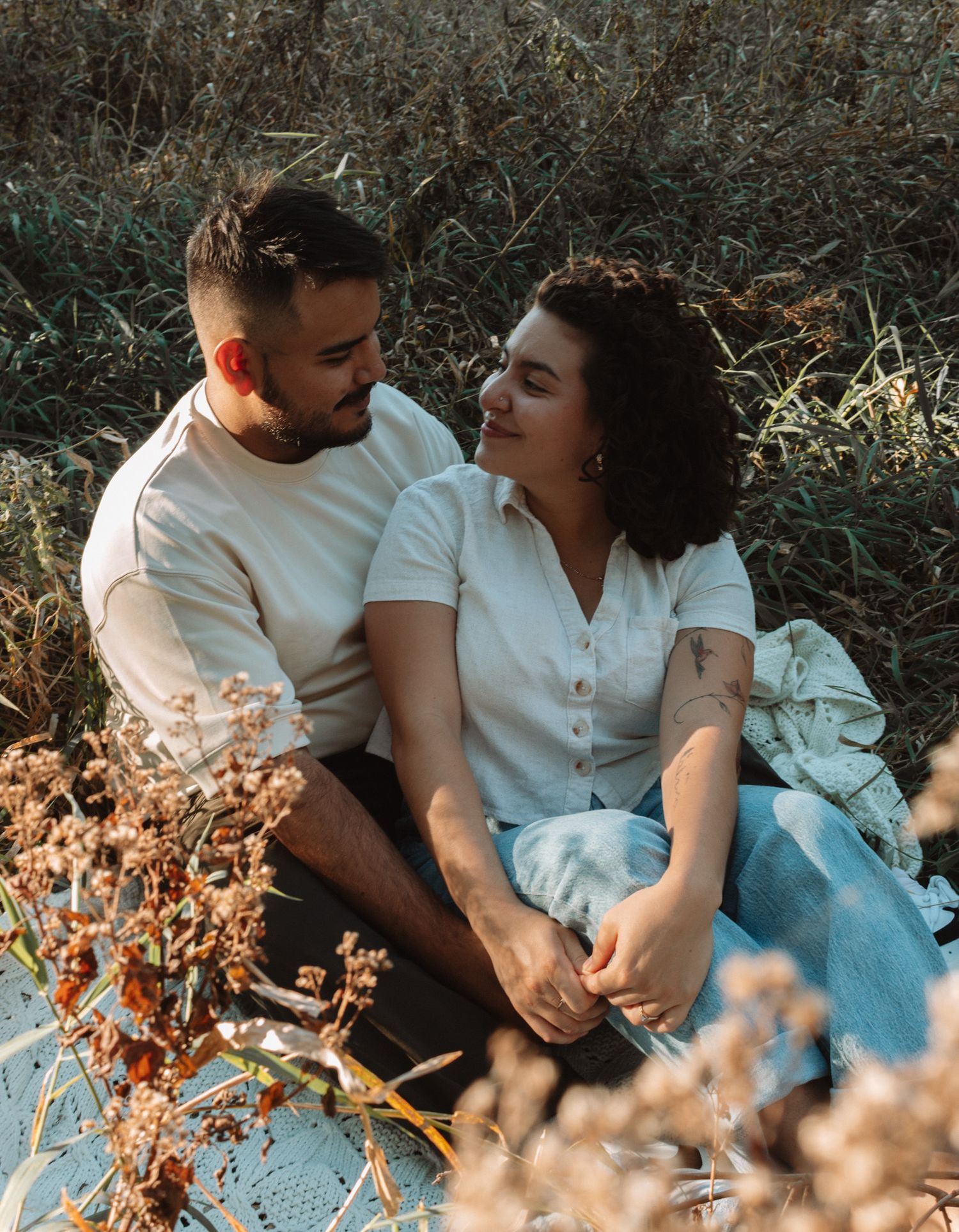 A couple lies together on a blanket outdoors surrounded by dried autumn foliage at sunset in a romantic setting.