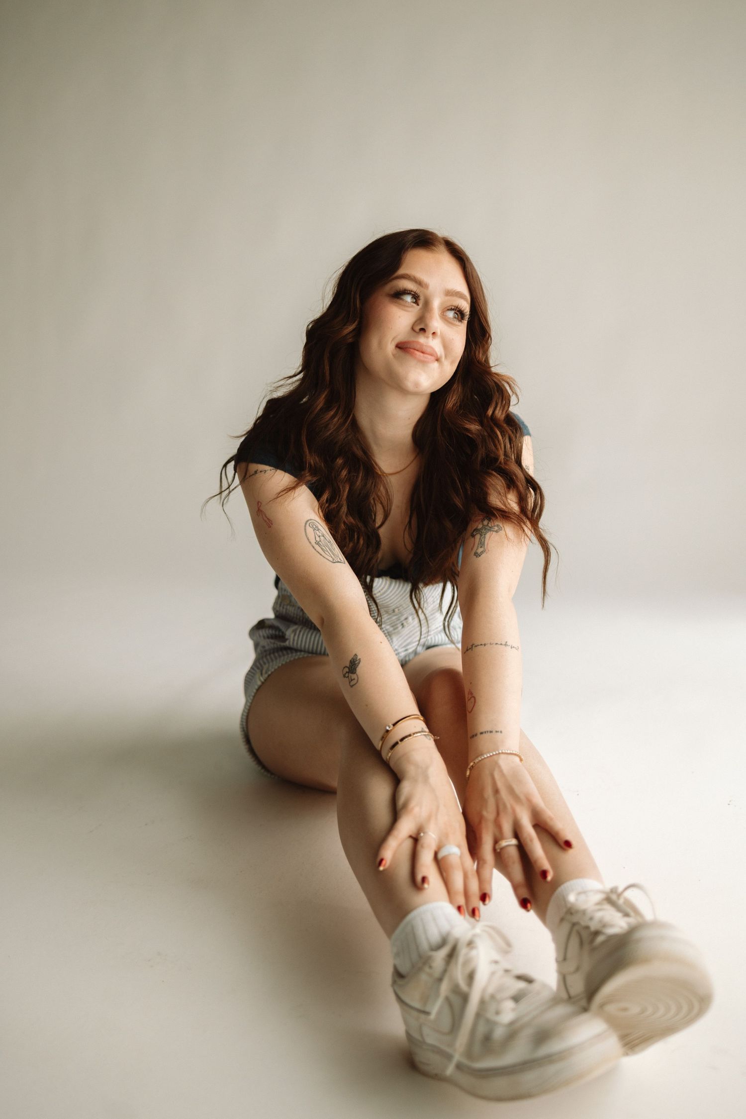 Portrait on white background wearing denim shorts and white sneakers with casual pose sitting on floor.