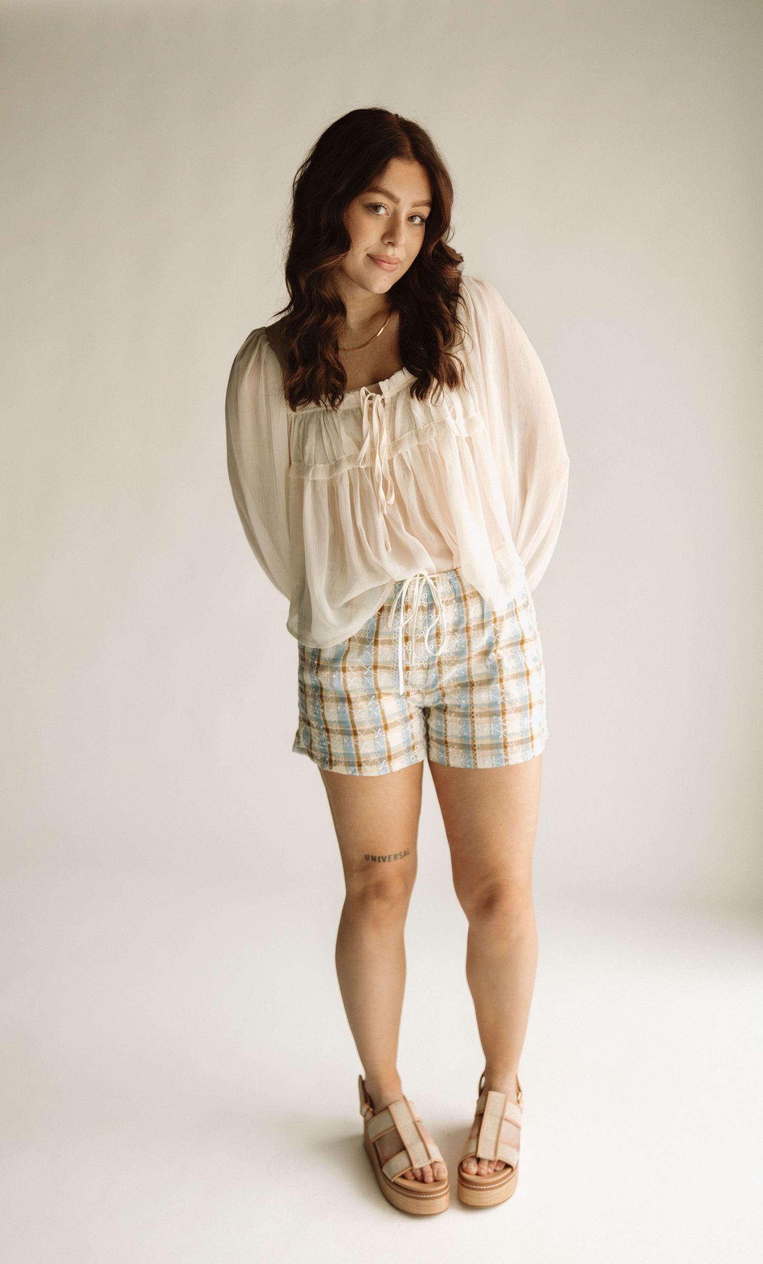 Model wearing a white peasant blouse with plaid shorts and platform sandals poses against a light backdrop.