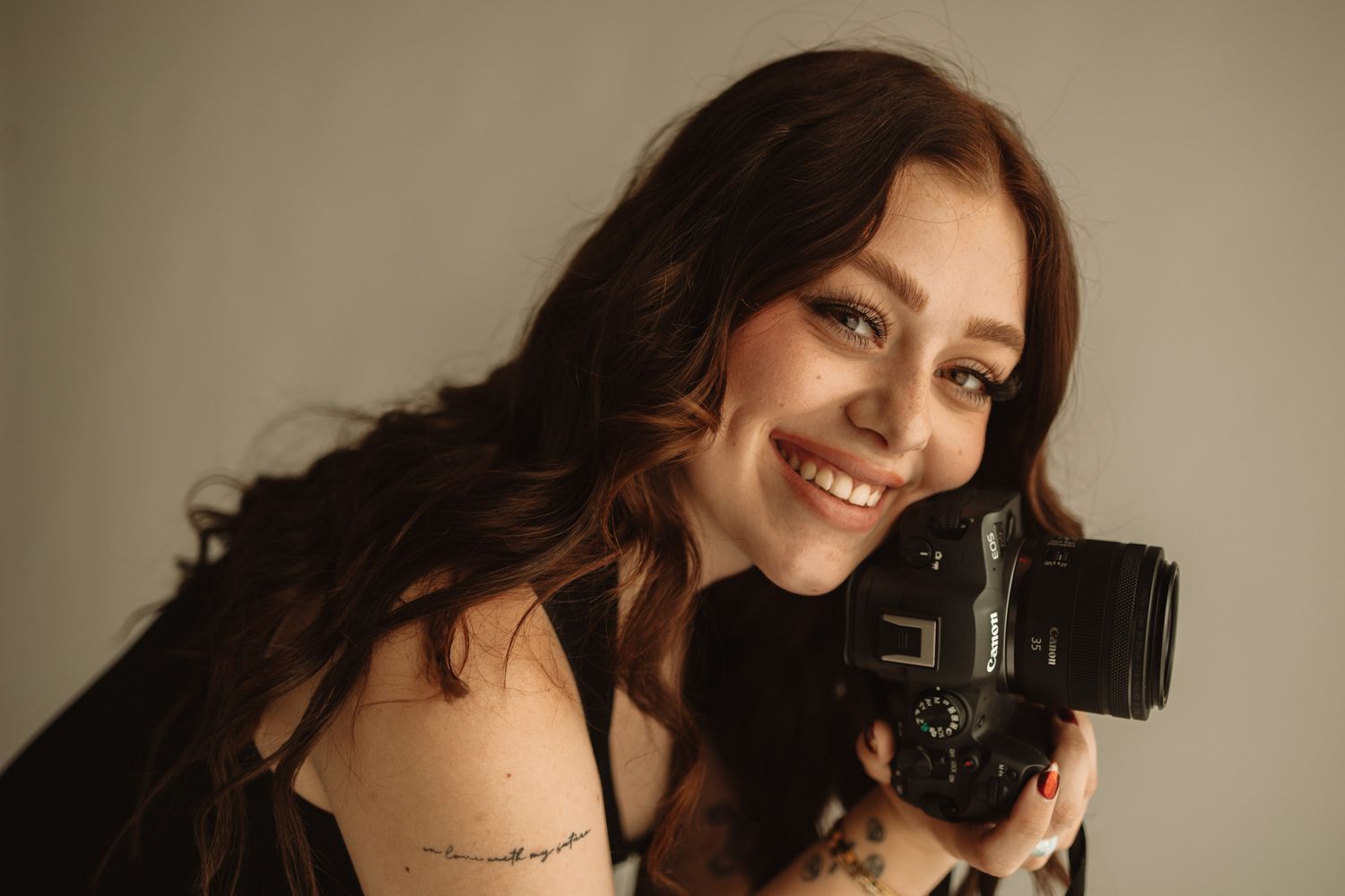 A professional photographer with dark curly hair smiles while holding a DSLR camera in a studio setting.