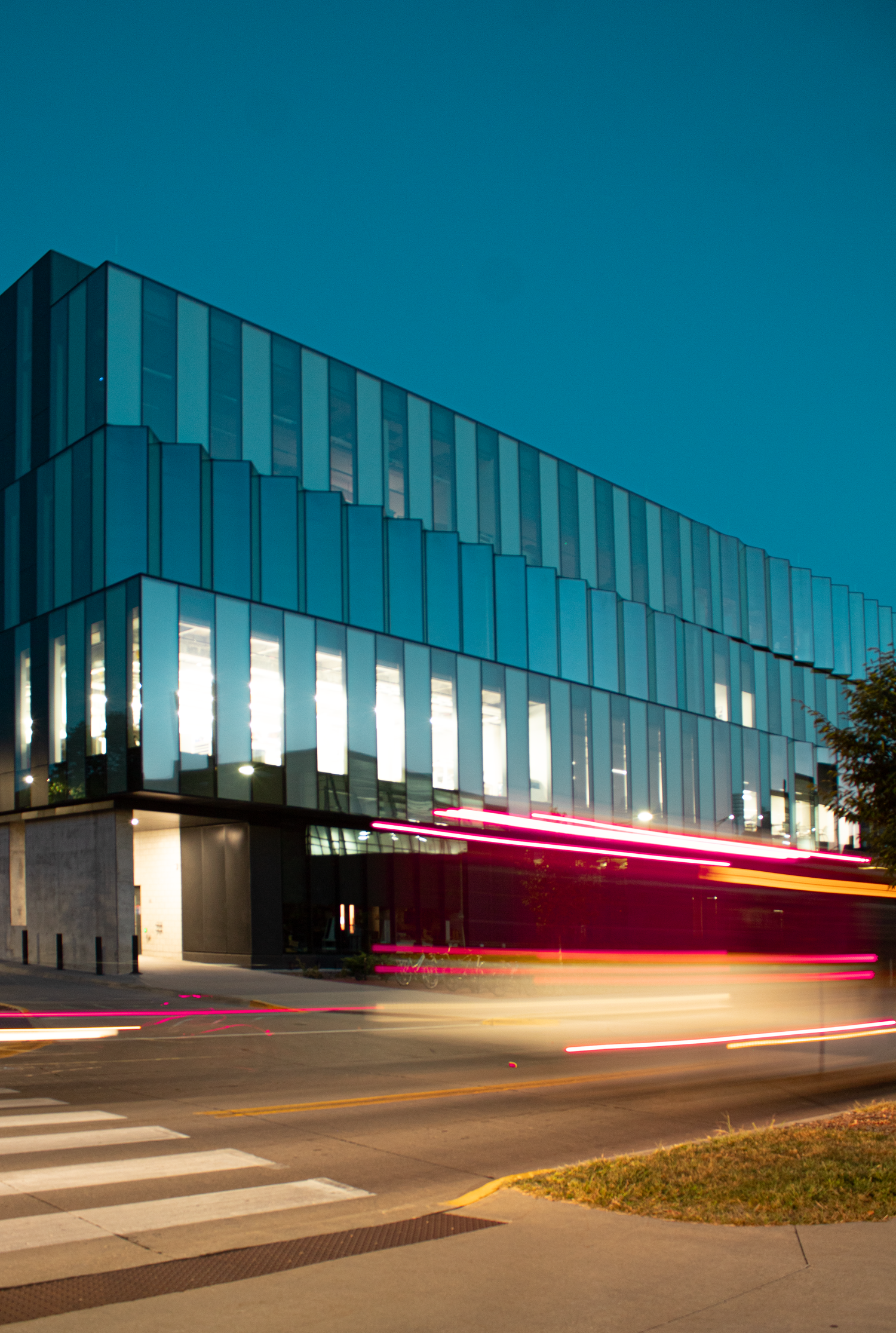 Light trails from passing vehicles create streaks across a modern glass building facade at dusk against a deep blue sky.